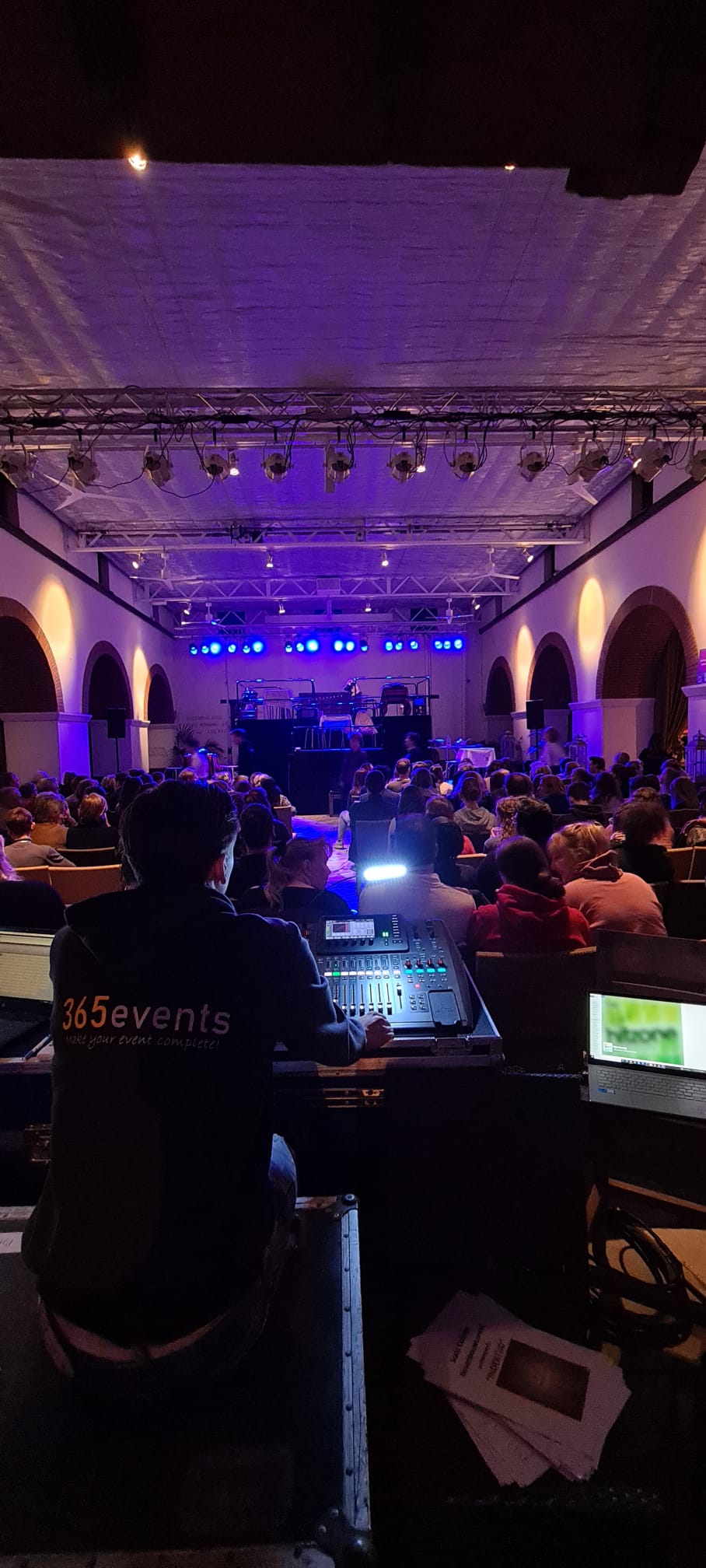Geluidstechnicus bij een concert, podiumverlichting die de band en het publiek in een grote zaal verlicht.