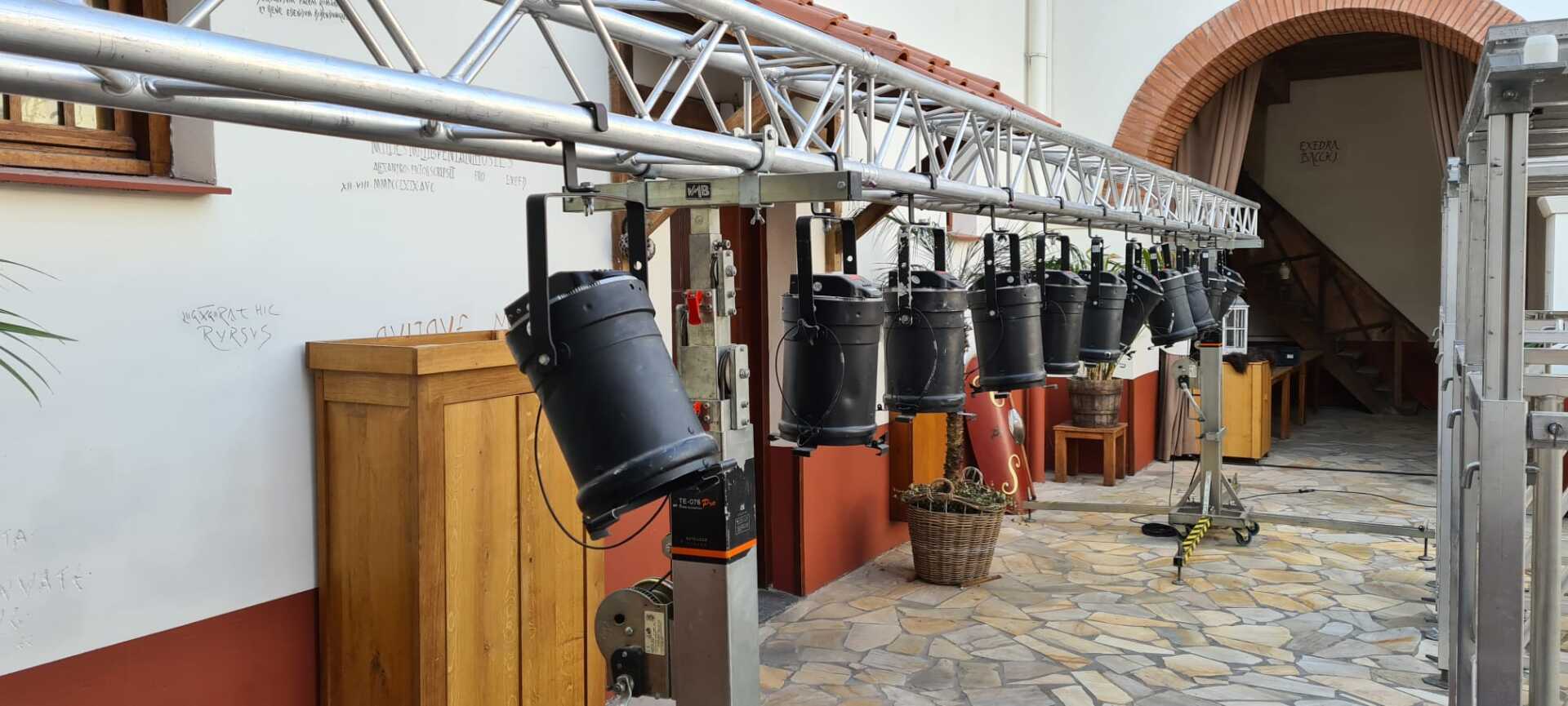Een rij zwarte podiumlampen hangt aan een metalen truss in een openluchtbinnenplaats.
