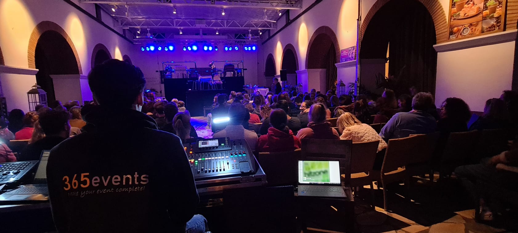 Een persoon bedient de geluidsmixer tijdens een concert in een grote zaal, met een publiek en podiumverlichting in het zicht.