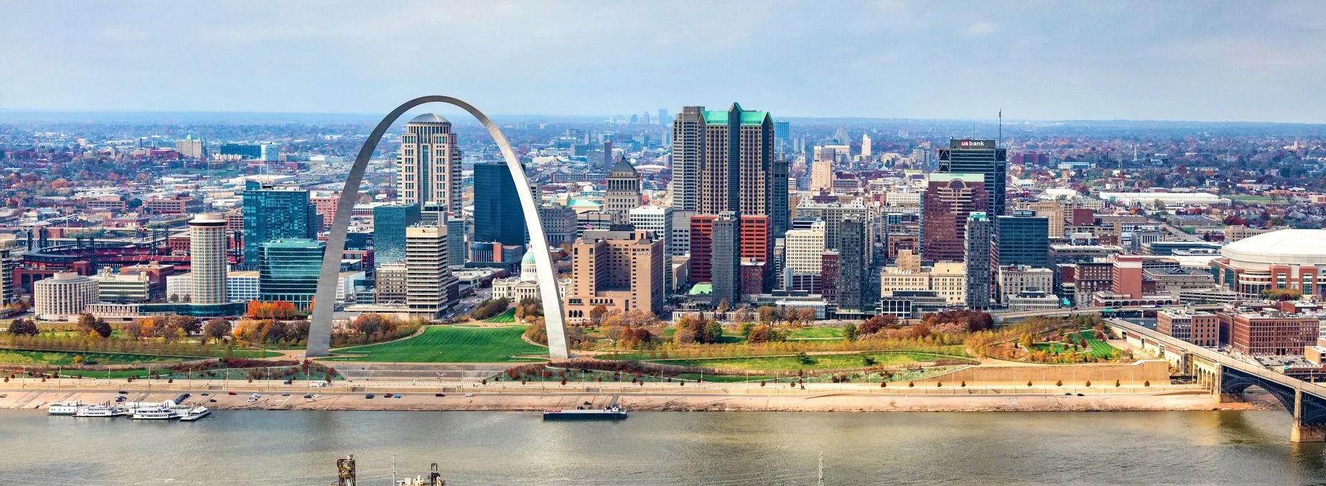 The Gateway Arch and the St. Louis skyline, seen from across a wide river. A bridge is on the right, and boats are near the shore.