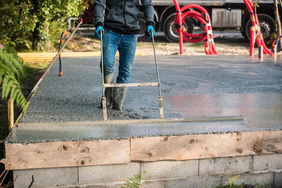 Person smoothing wet concrete on a driveway with a trowel, near a garage and red chairs.
