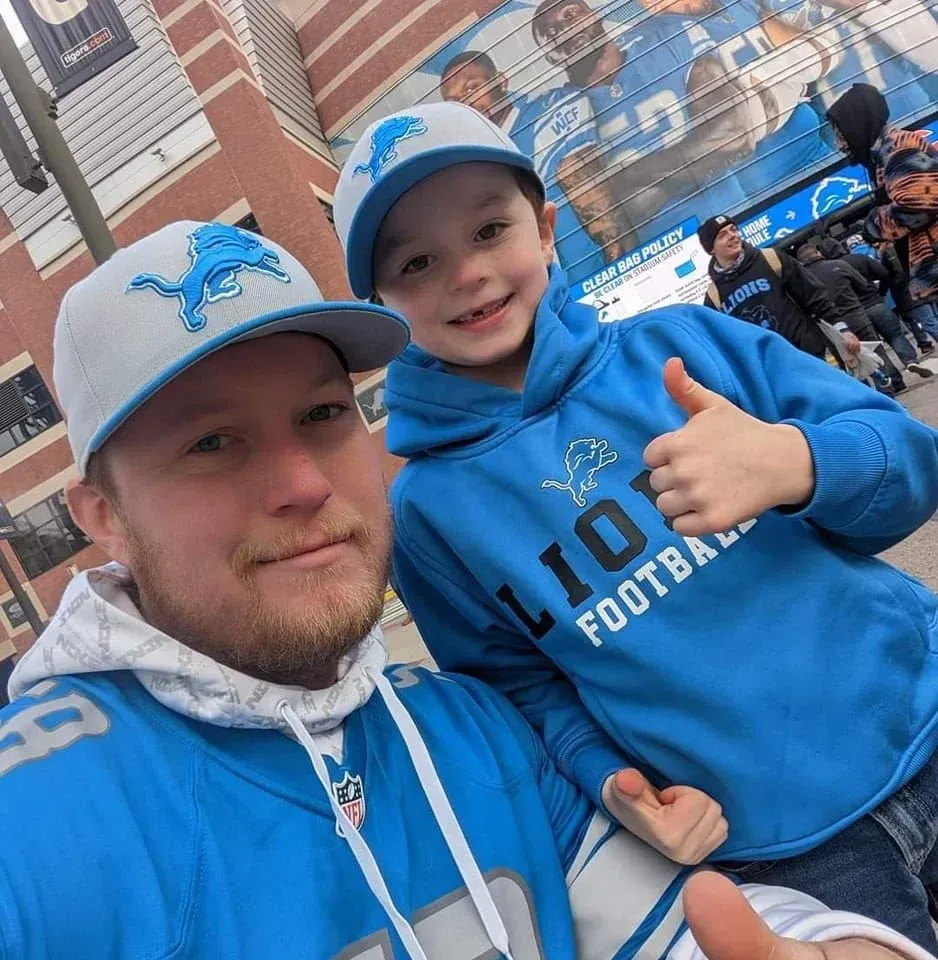 Two Detroit Lions fans in blue gear smiling at a football game, one giving a thumbs-up