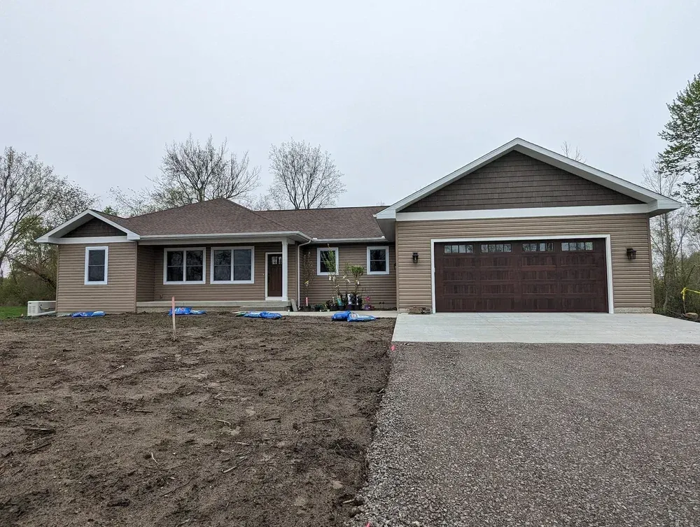Single-story tan house with brown garage door, gravel driveway, and bare front yard under overcast sky