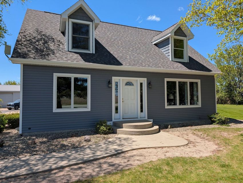 Blue house with white trim, steep roof, front door, and dormer windows on a gravel driveway under a clear sky