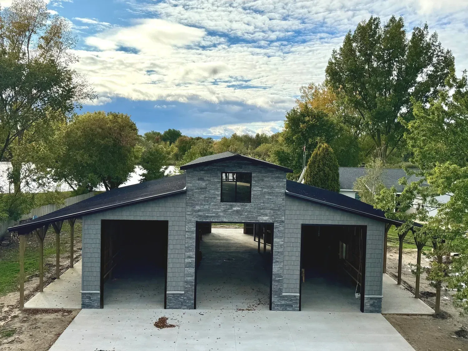 Gray triple-bay garage with open doors, surrounded by trees under a cloudy sky