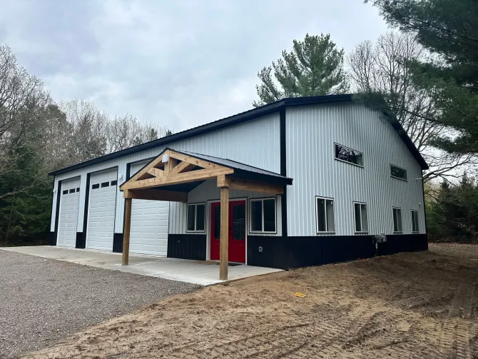 White metal barn-style building with red doors and covered entry on a gravel lot, surrounded by trees