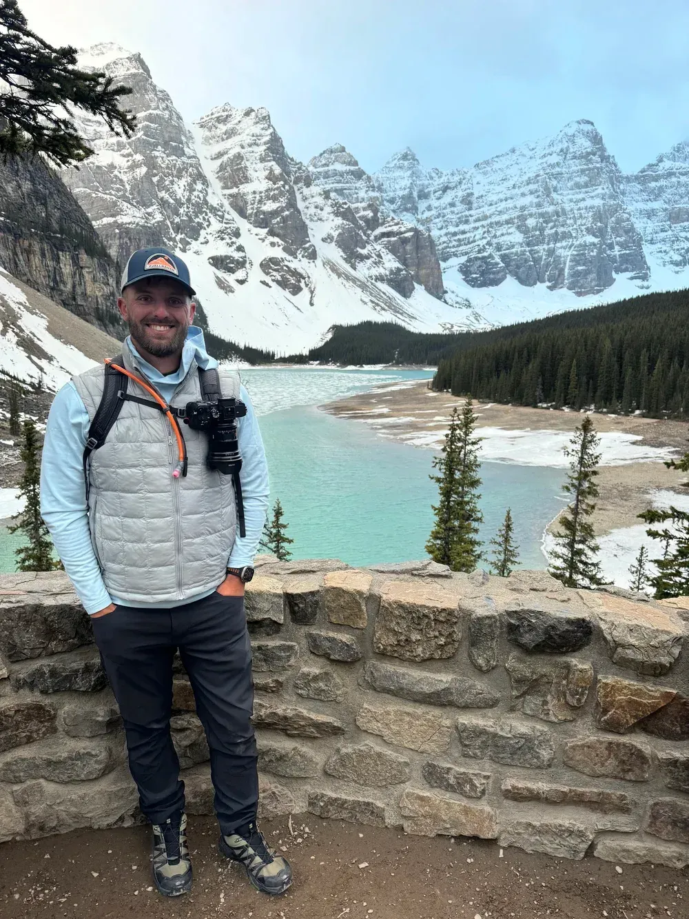 Smiling hiker in a gray jacket stands by a stone wall with turquoise lake and snowy mountains behind.