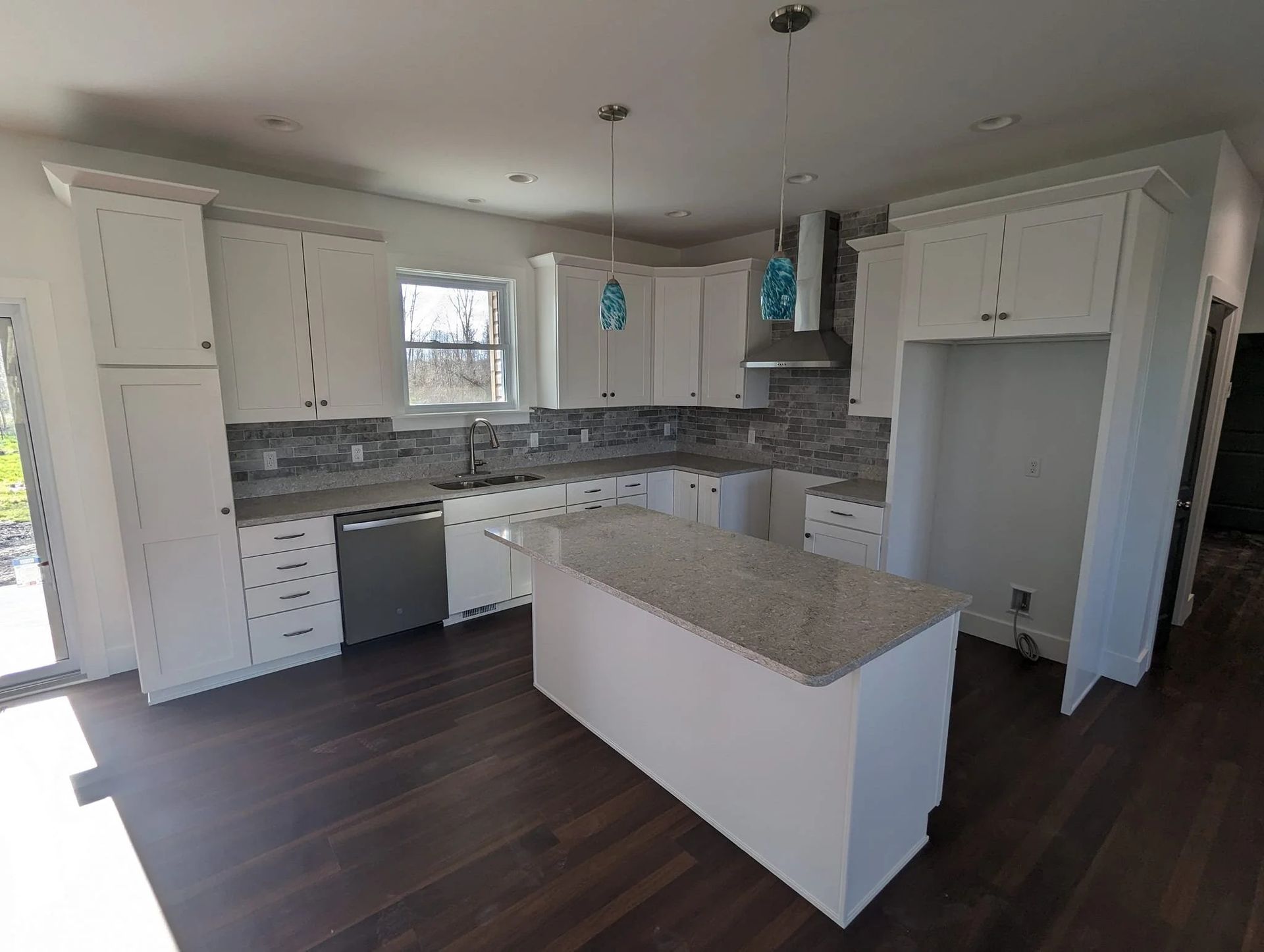 Modern white kitchen with dark wood floors, gray tile backsplash, and a granite-topped island.