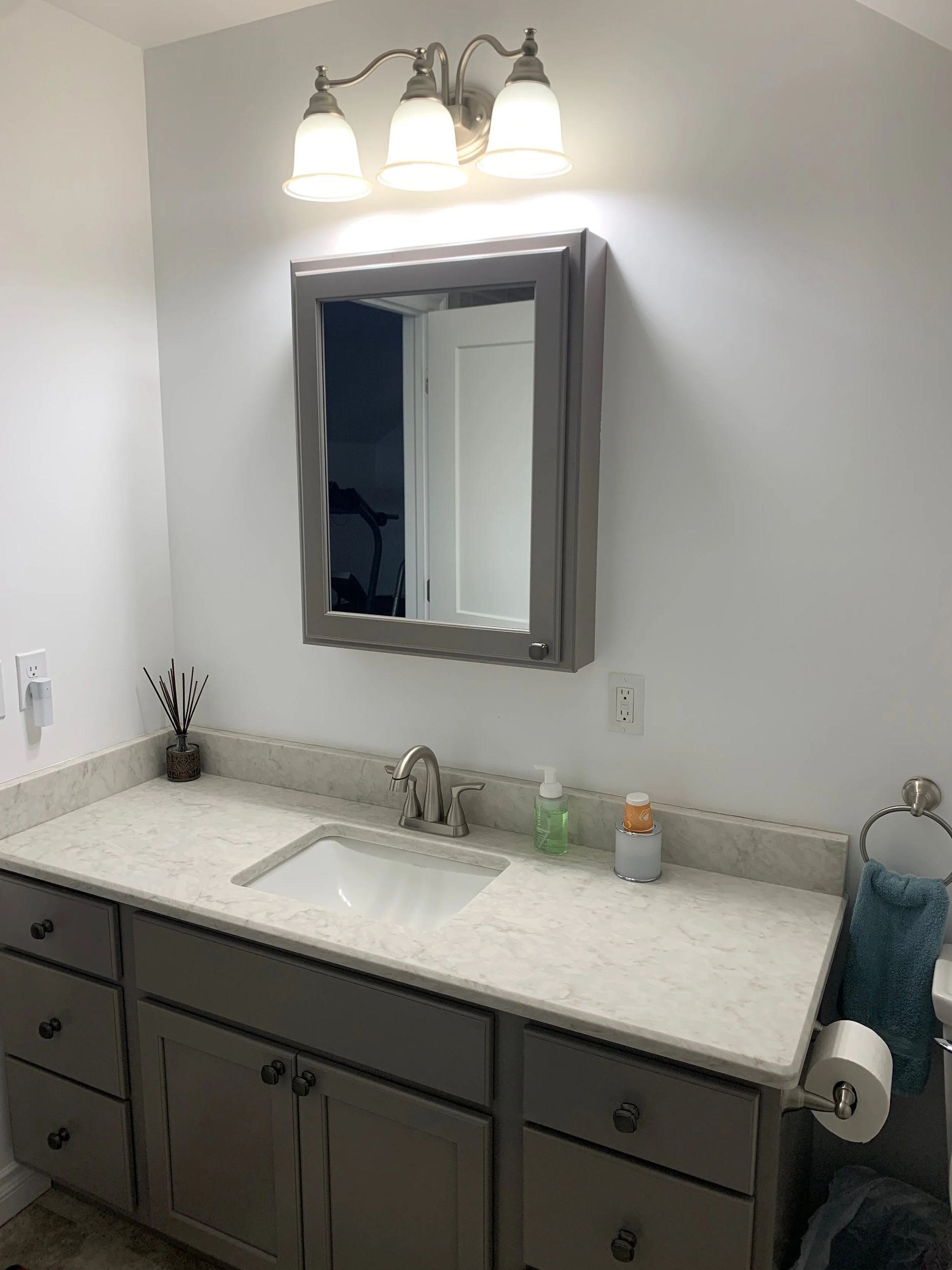 Bathroom vanity with gray cabinets, white countertop, mirror, and three-light fixture above sink.