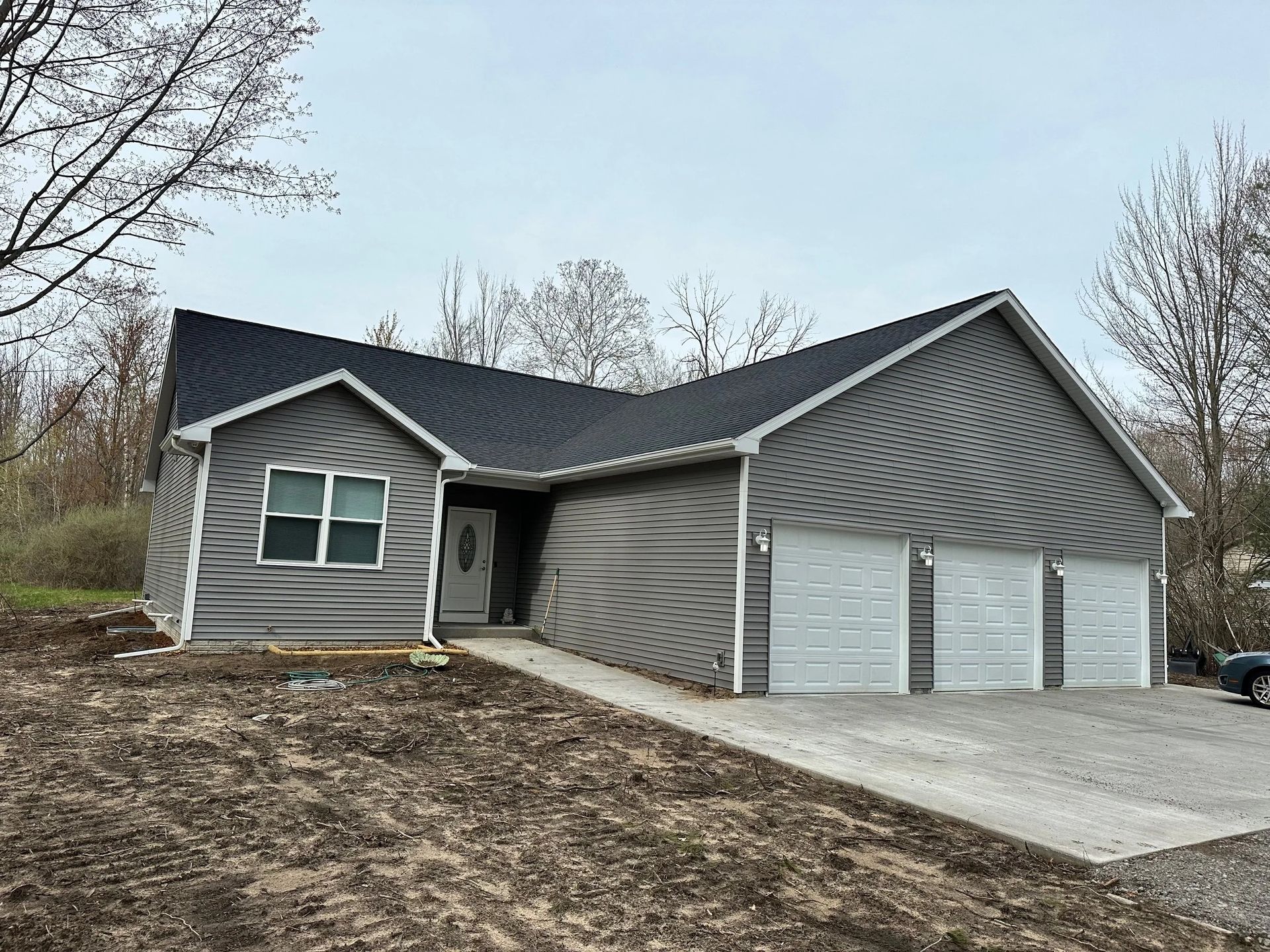 Gray house with warped siding, three garage doors, and a concrete driveway on a wooded lot