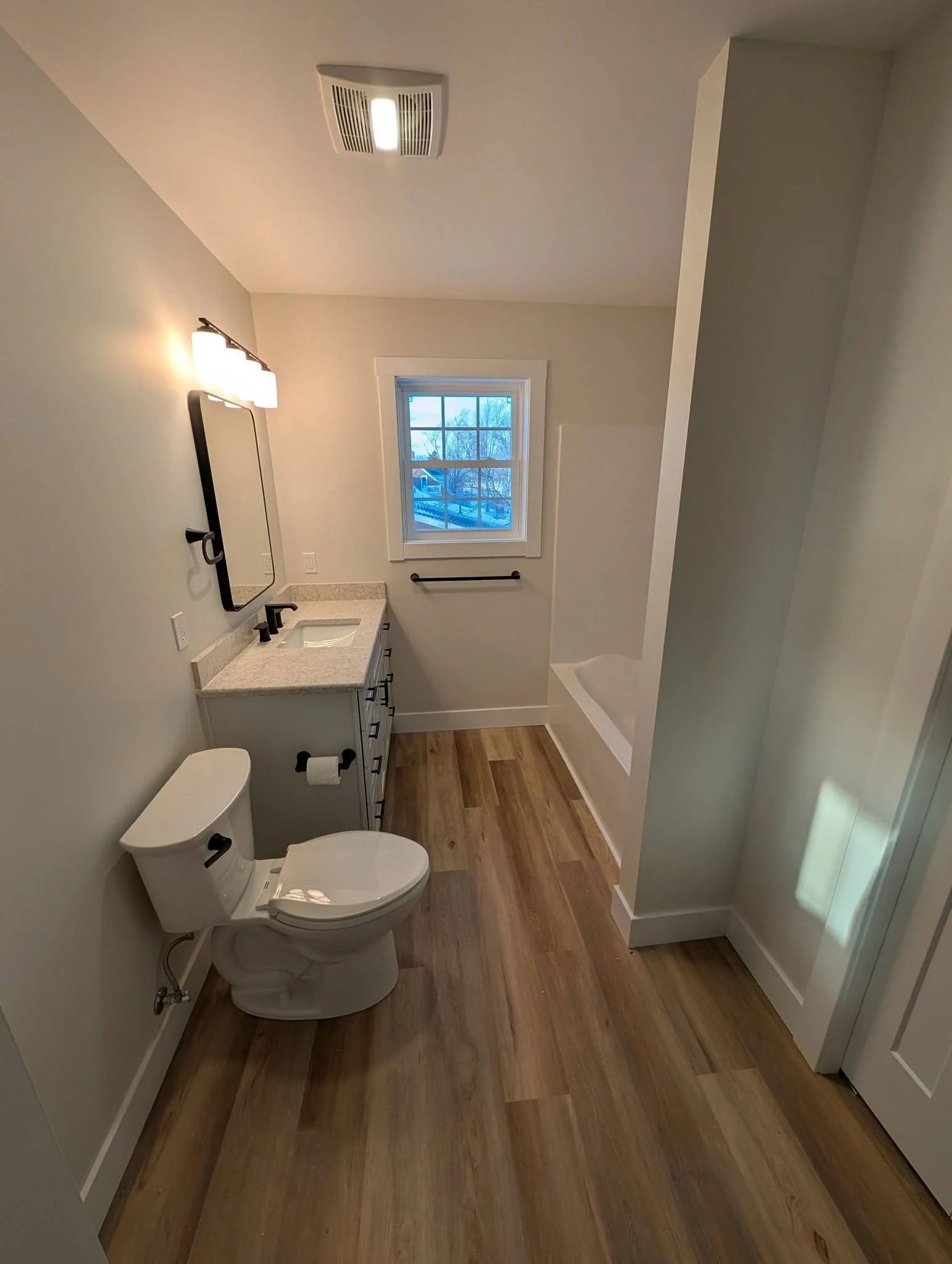 Narrow bathroom with toilet, sink, mirror lights, wood-look floor, and window at the end.
