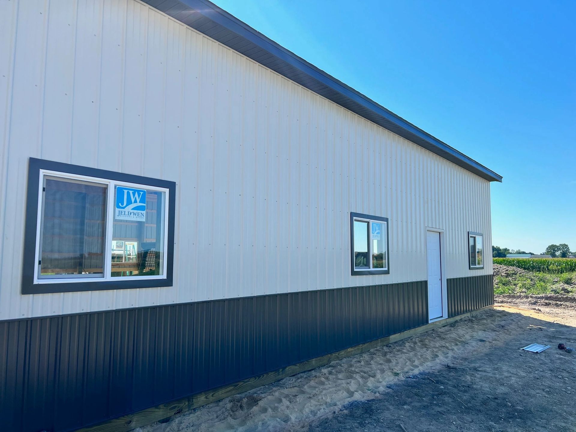 White metal building with black trim, windows, and a side door on a dirt lot under a clear blue sky
