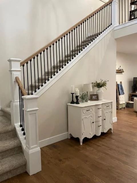 White staircase with dark railing beside a small white cabinet on wood flooring in a bright home interior