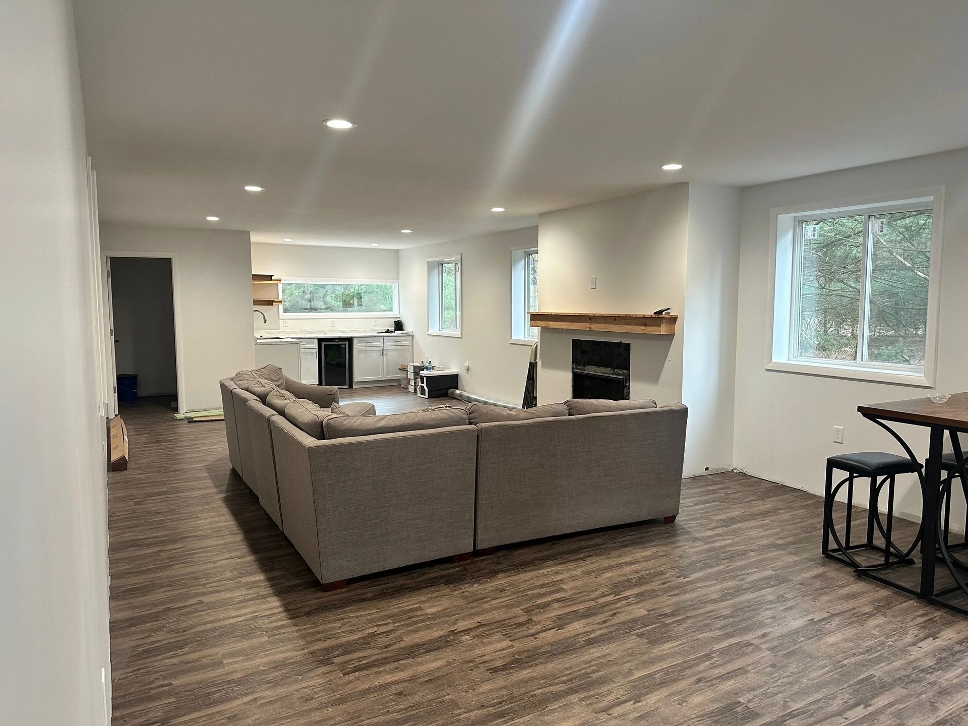 Open-concept living room with gray sectional, fireplace, windows, and kitchen area in the background