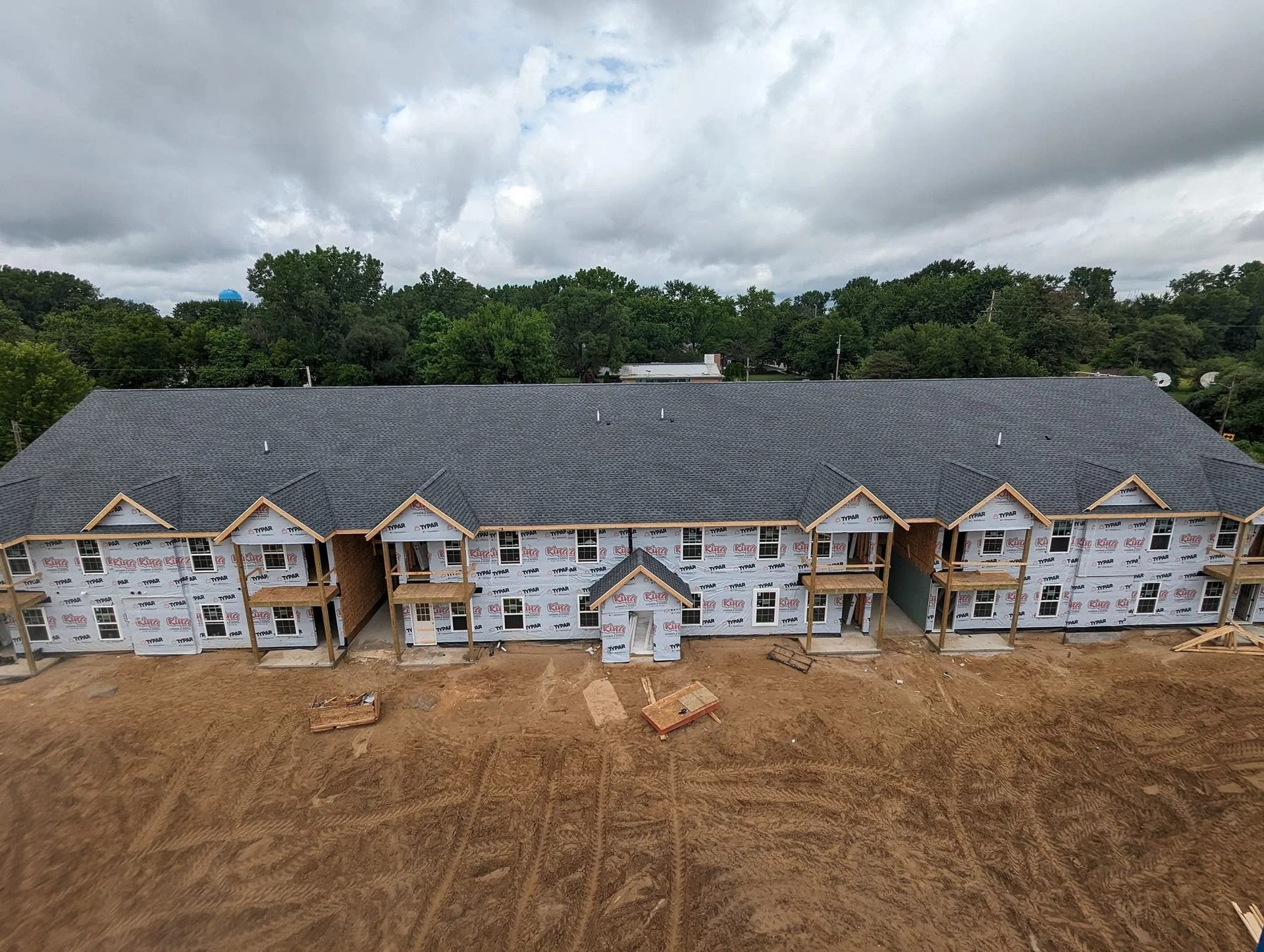 Large multi-unit house under construction with unfinished white siding, gray roof, and dirt lot in front.
