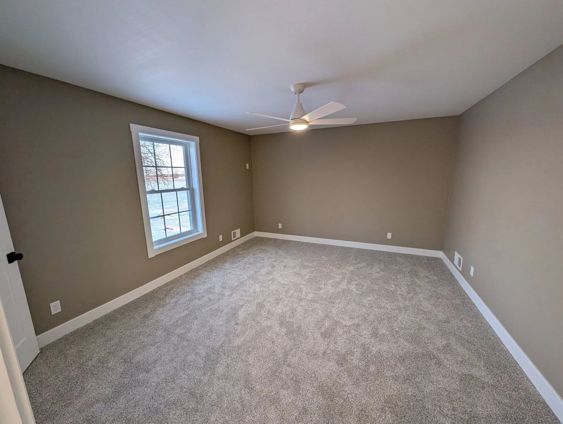 Empty beige bedroom with carpet, window, and ceiling fan on.