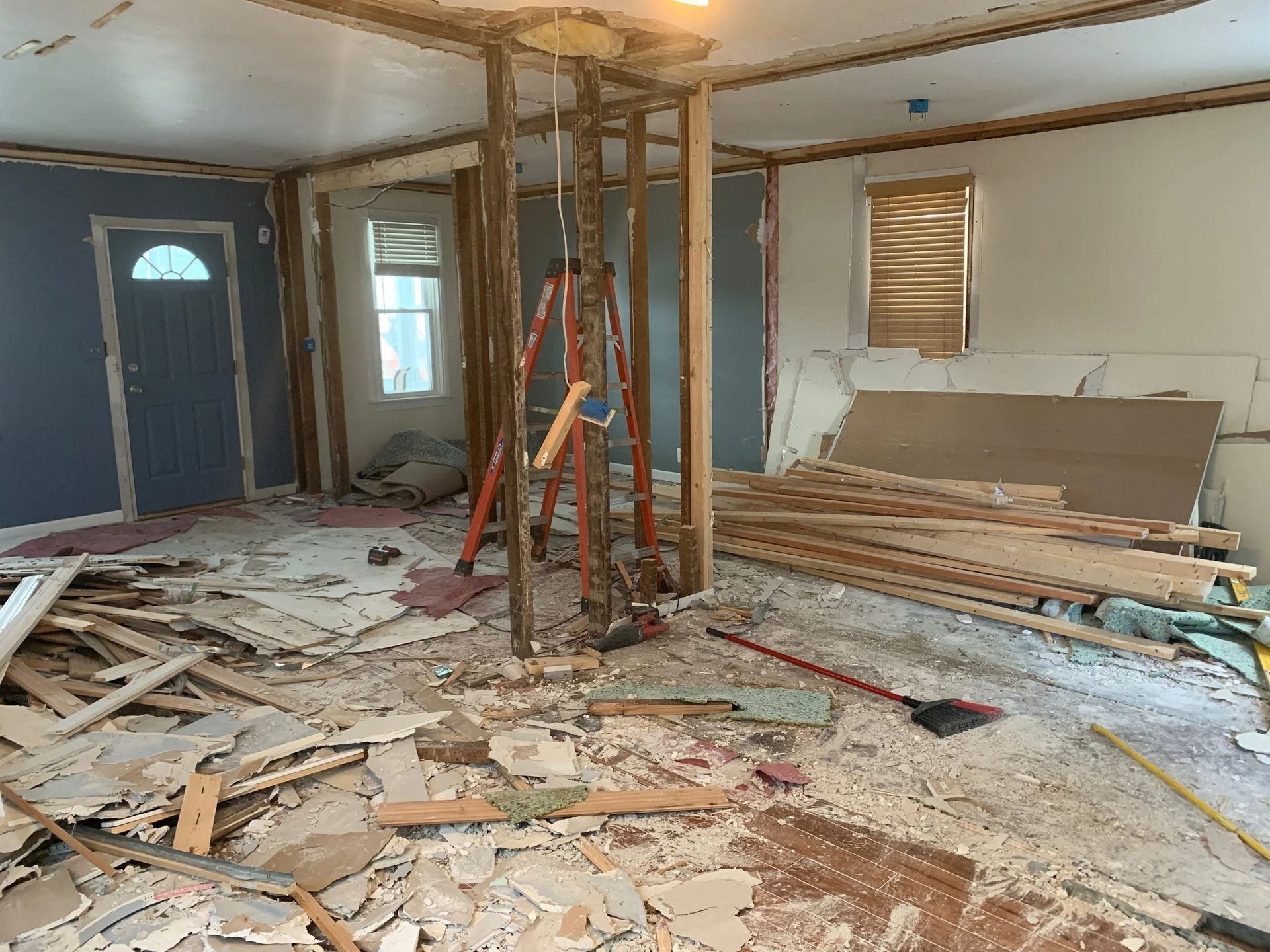 Partially demolished room with exposed studs, debris, ladders, and stacked lumber during renovation