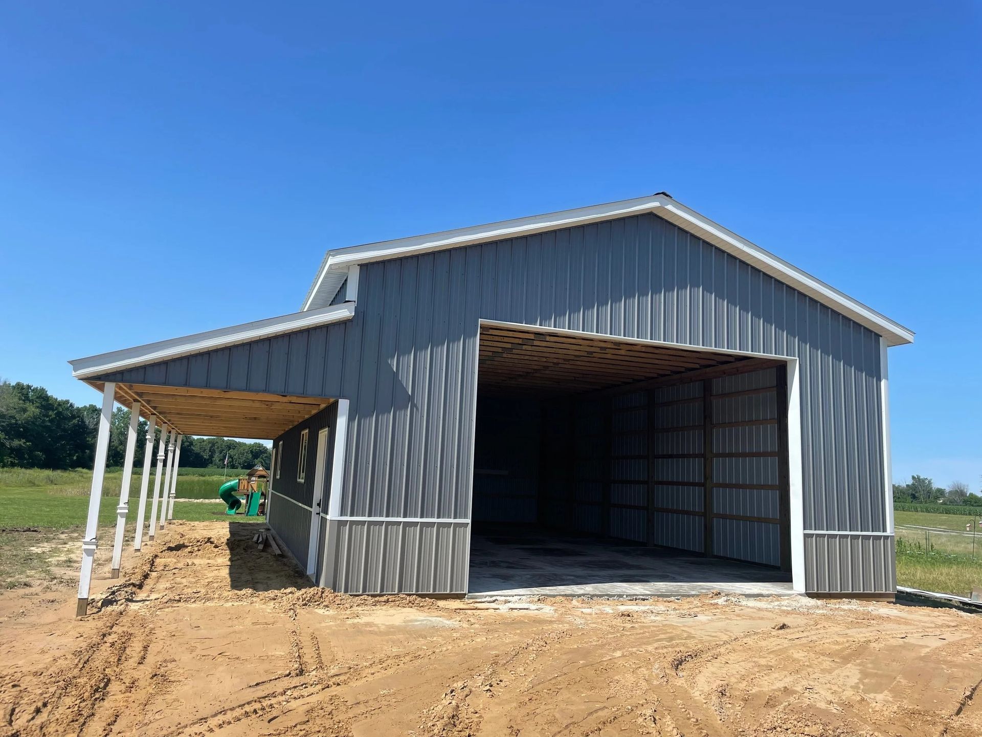 Gray metal barn under construction on a dirt lot, with a covered side bay and open front entrance.