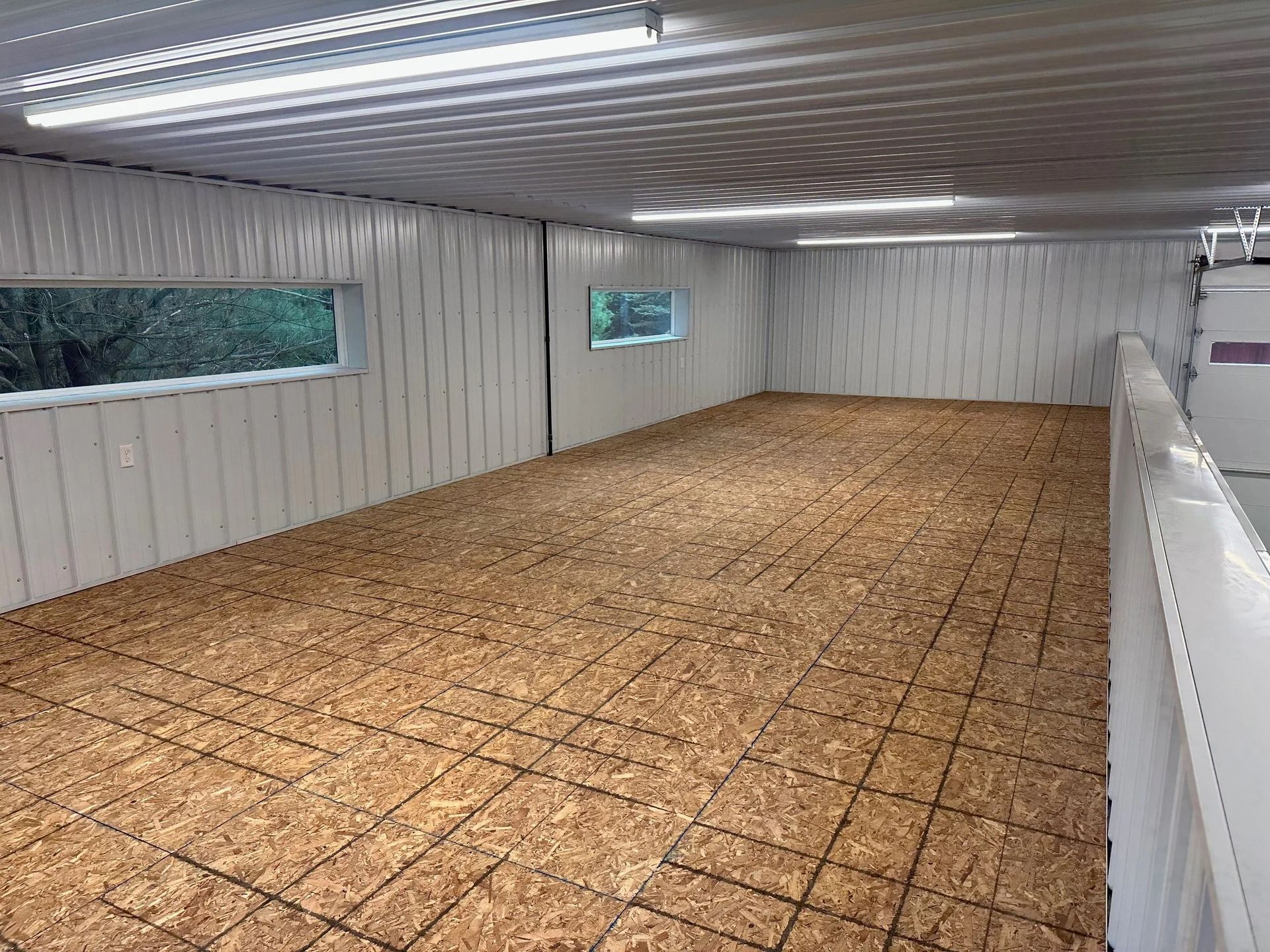 Empty indoor room with white walls, fluorescent lights, and a plywood floor.