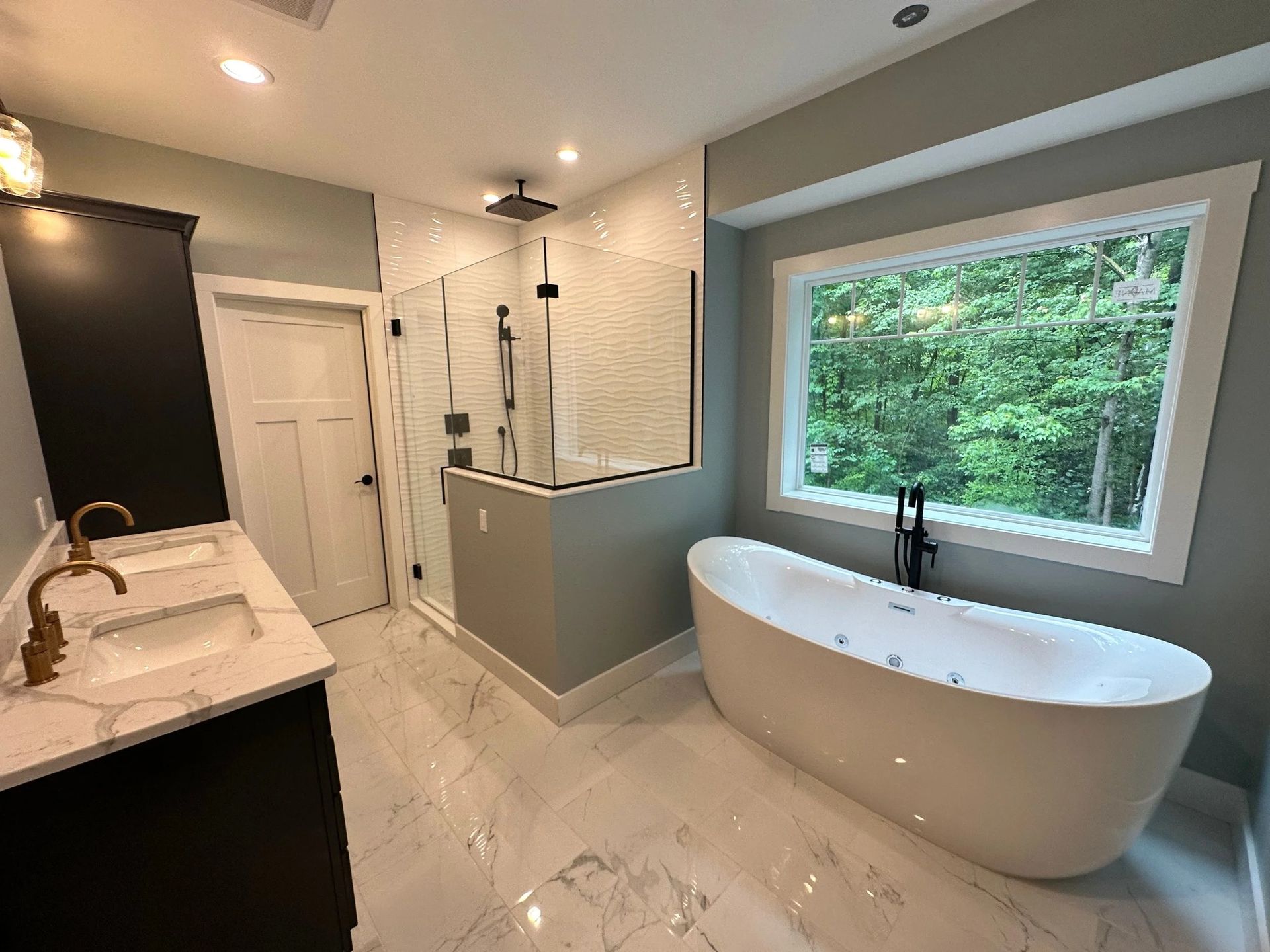 Modern bathroom with freestanding tub, glass shower, white marble tile, and large window overlooking greenery