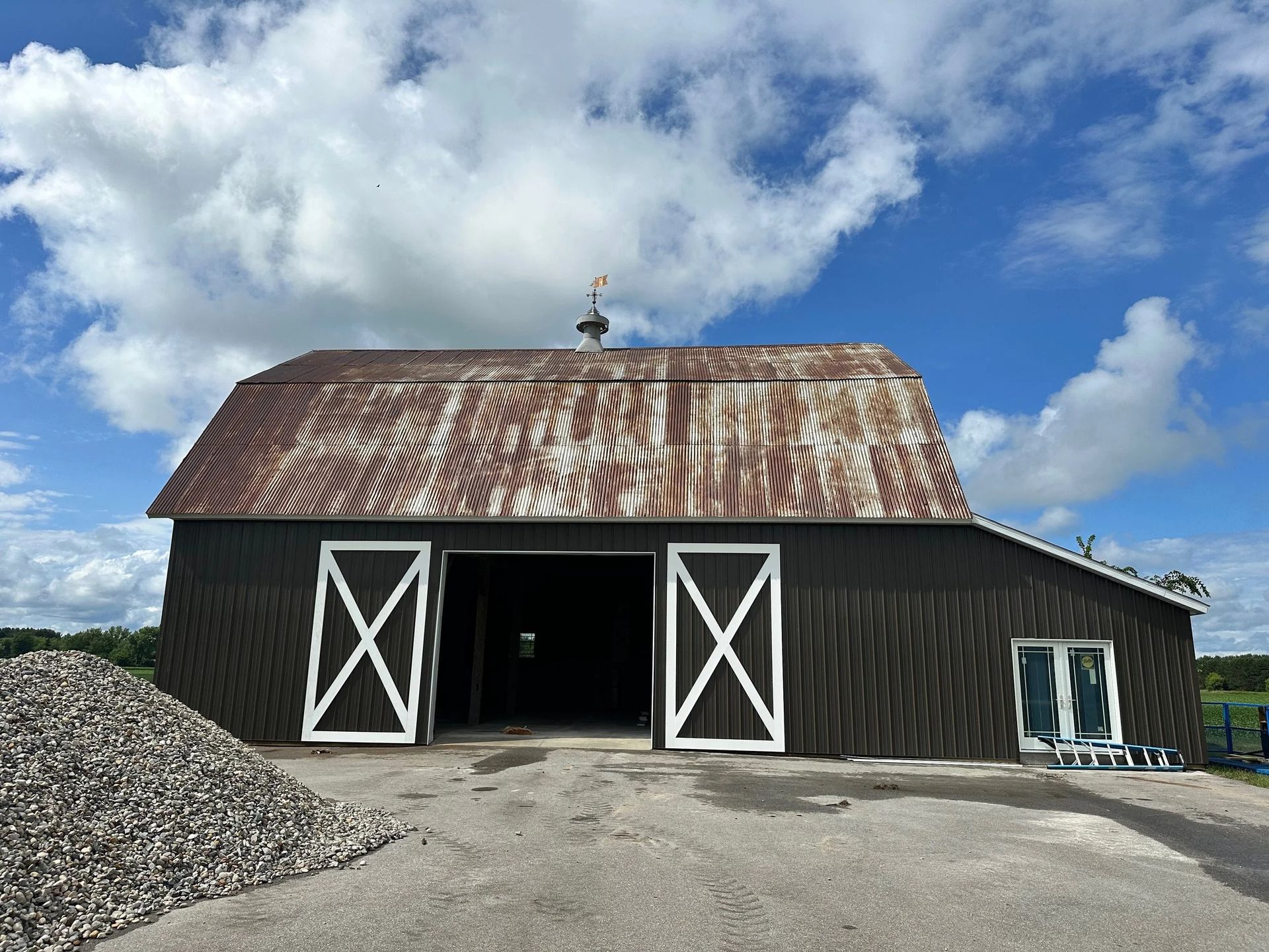 Barn with rusted metal roof, open white doors, and gravel driveway under a cloudy sky