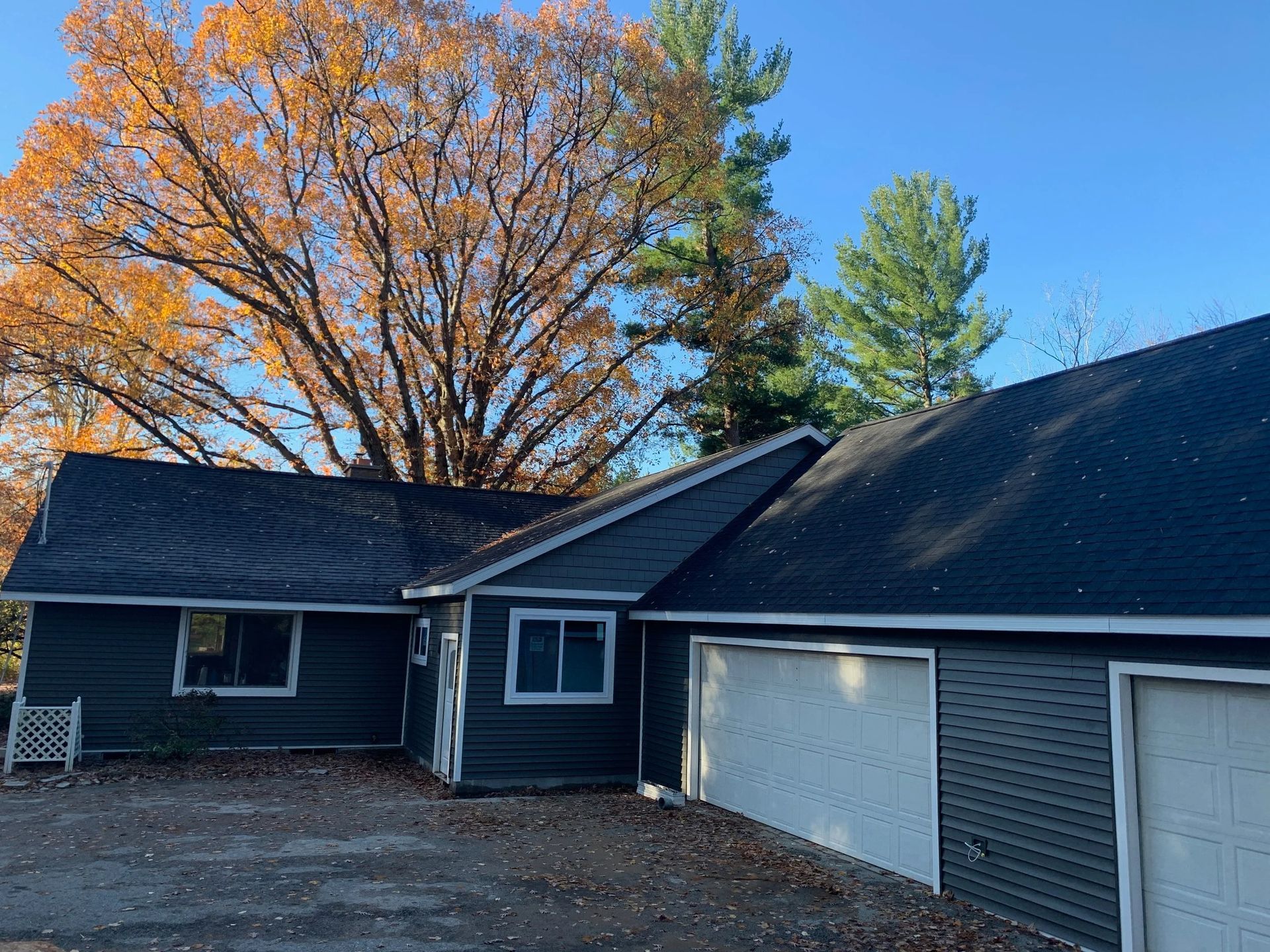 Suburban house with dark siding, white garage doors, and autumn trees under a clear blue sky