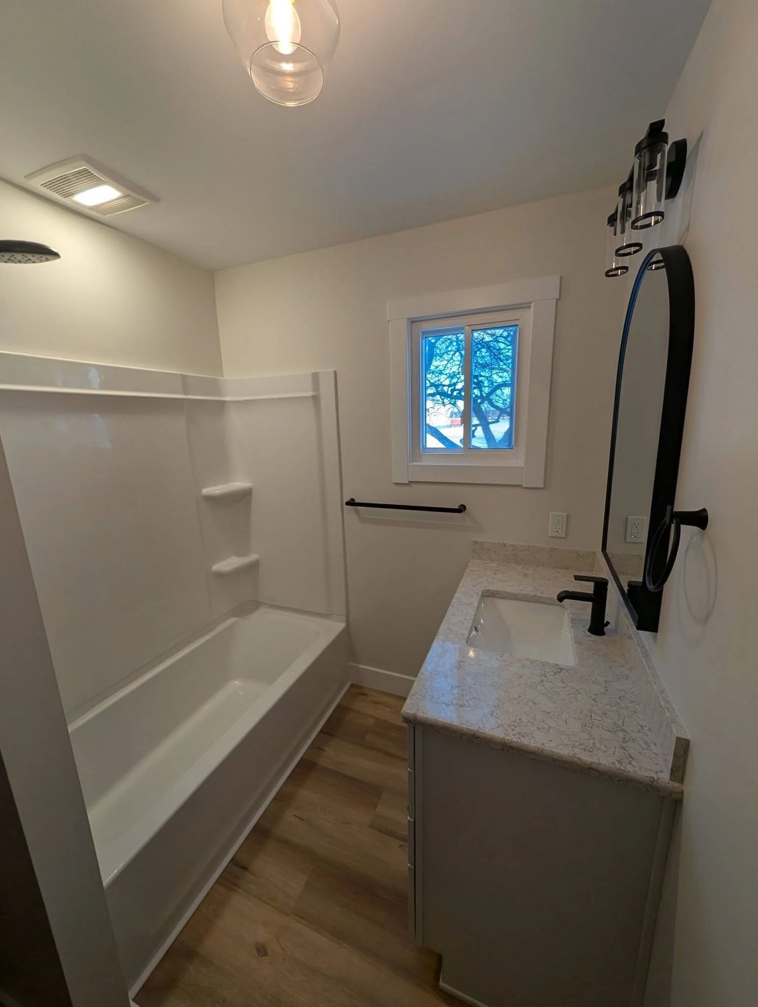 Small bathroom with white tub, floating shelves, vanity, and a window.