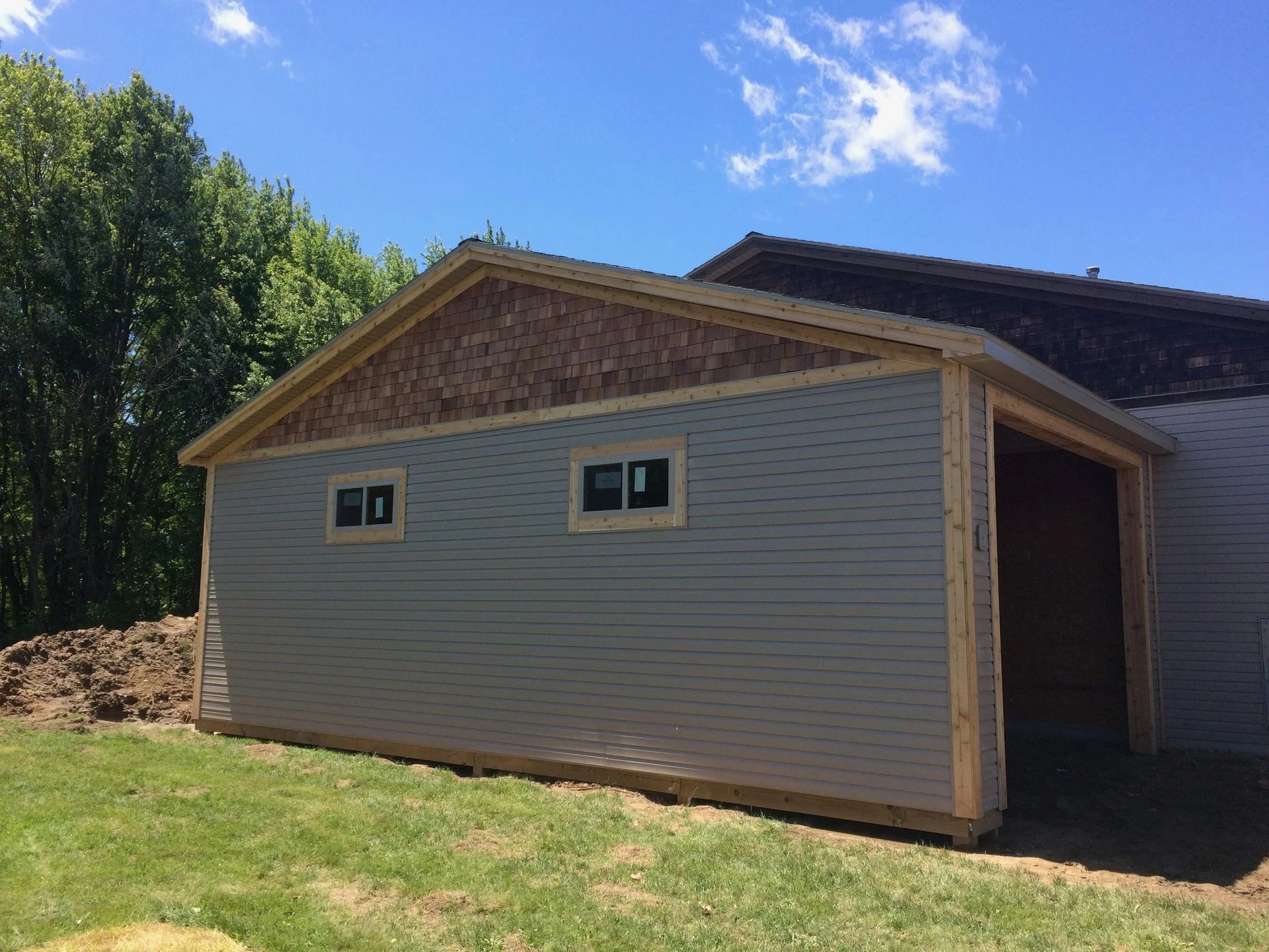 Small gray shed with white trim and brown roof on a grassy lot under a blue sky.