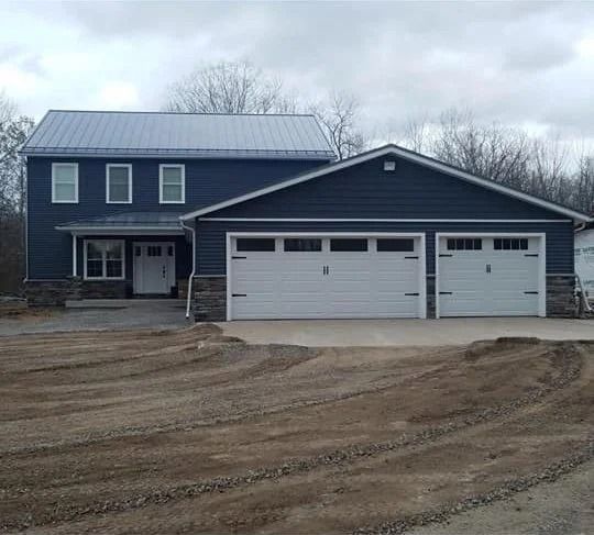 Two-story dark blue house with white trim and a three-car garage on a muddy lot under cloudy skies
