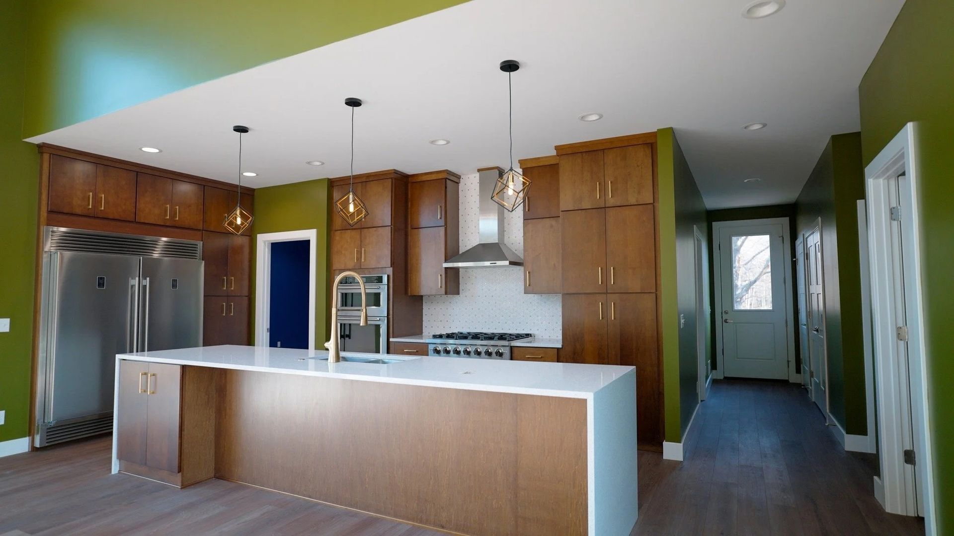 Modern kitchen with wood cabinets, white island, stainless fridge, and green walls, viewed from the dining area