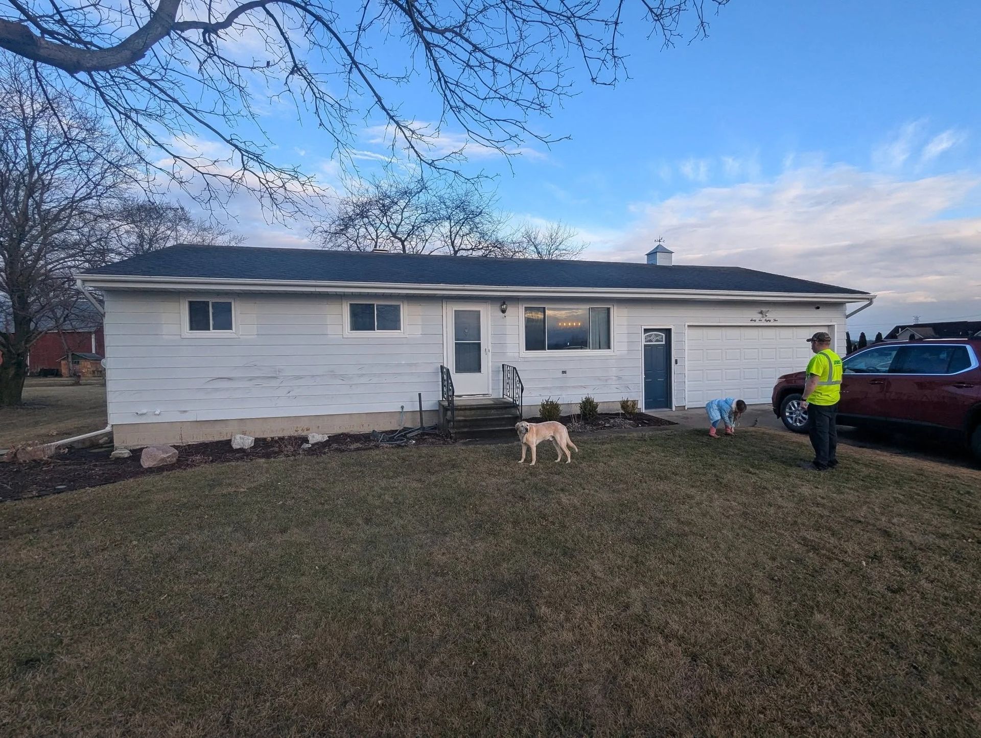 Single-story white house with a dog in the yard and a person near a red truck at dusk