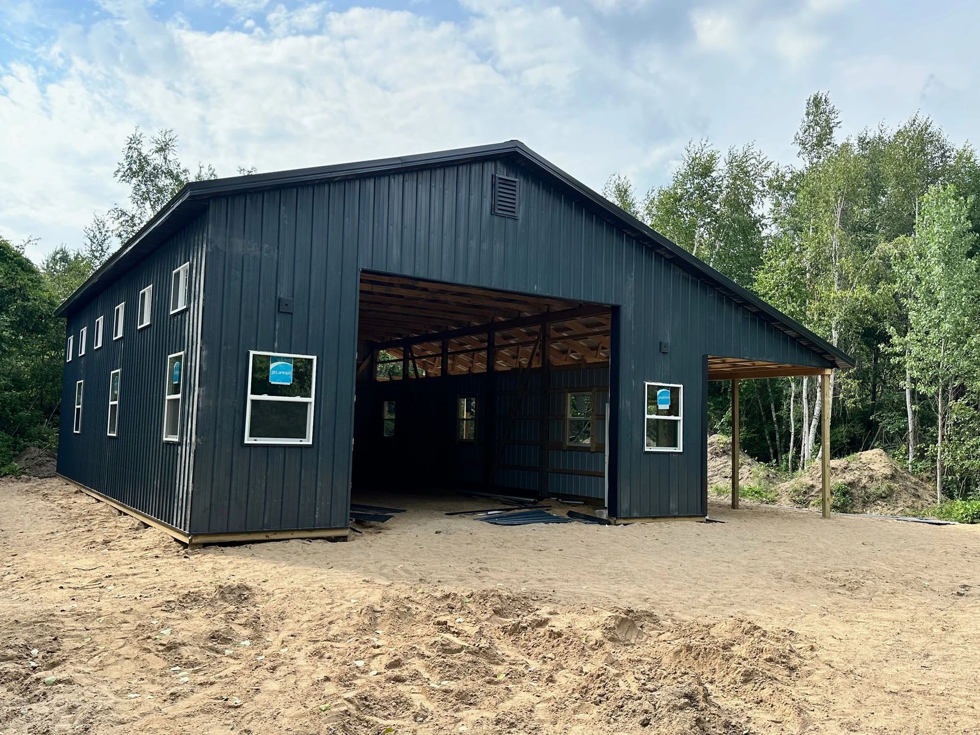 Black metal barn-like building with open front, windows, and sandy clearing surrounded by trees