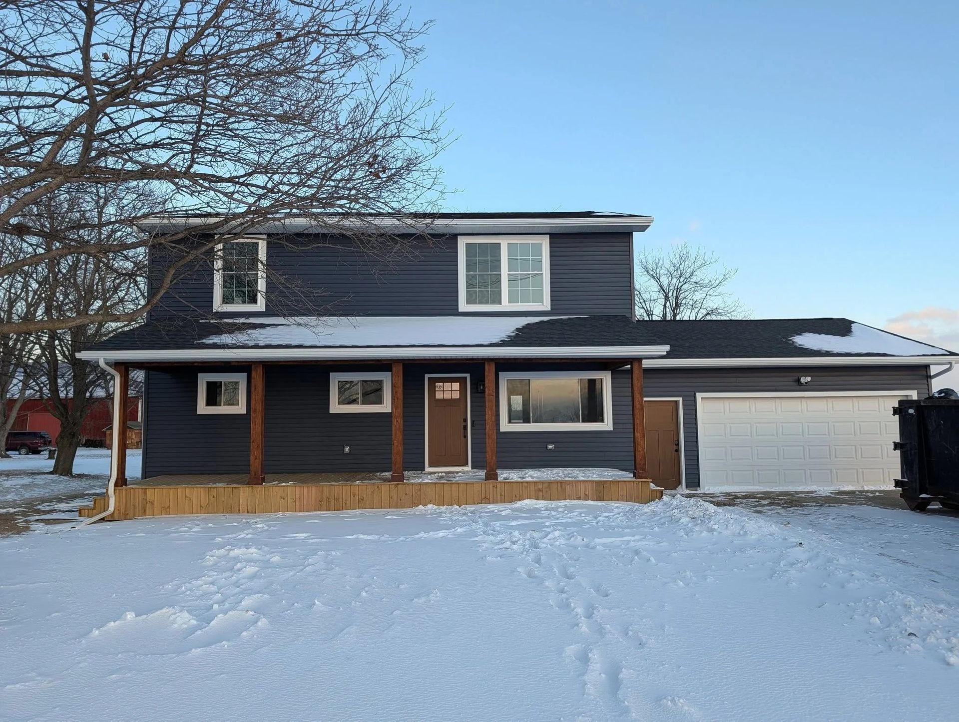Snow-covered two-story dark house with white garage and bare tree in front