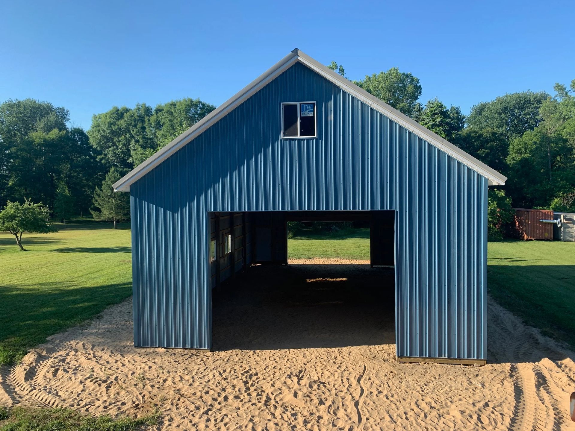 Blue metal barn with a large open doorway on a sandy lot, surrounded by trees and grass.