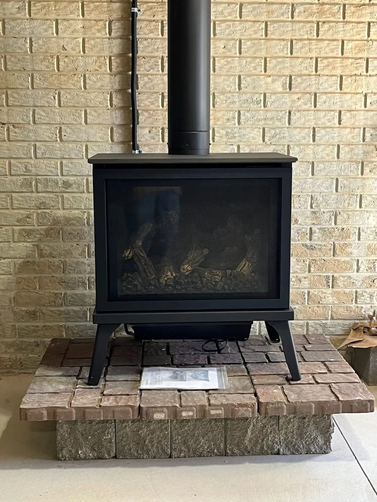 Black wood stove on a brick hearth against a beige brick wall, with a chimney pipe above.