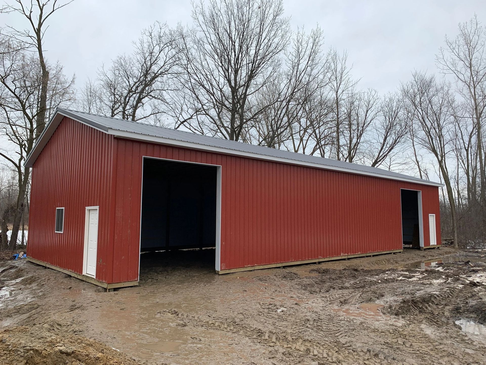 Red metal barn with open doors on muddy ground, leafless trees in the background