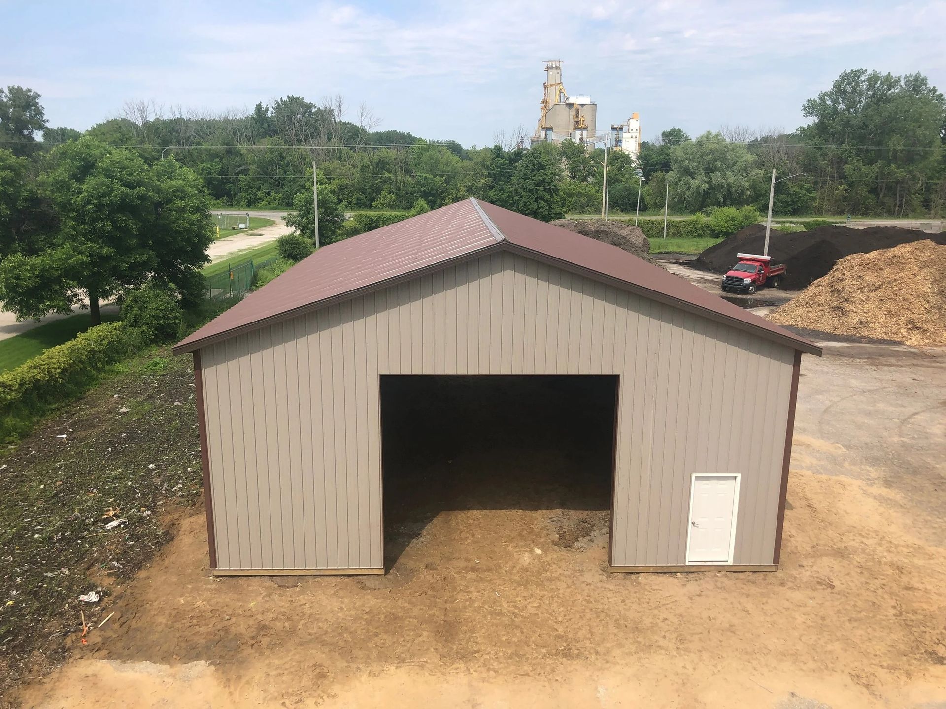 Small tan shed with a brown roof on a dirt lot, surrounded by trees and a rural landscape.