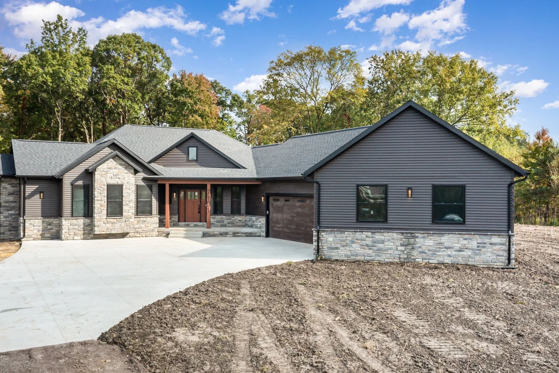 Modern gray single-story house with stone accents, dark garage, and large curved driveway under trees.