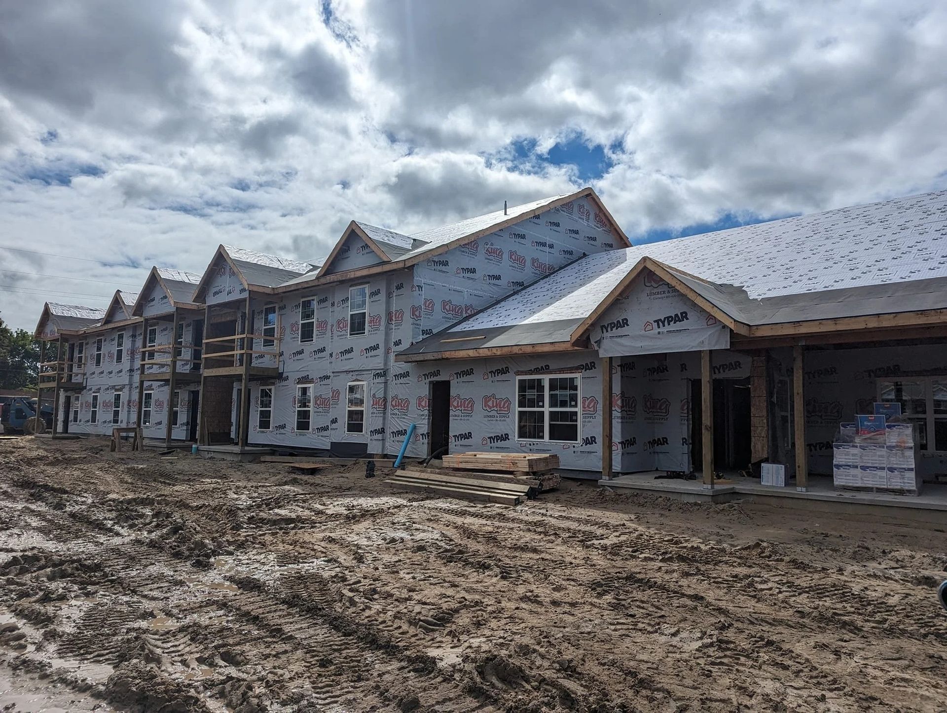 Partially built row of townhouses with exposed framing and muddy construction site under cloudy sky