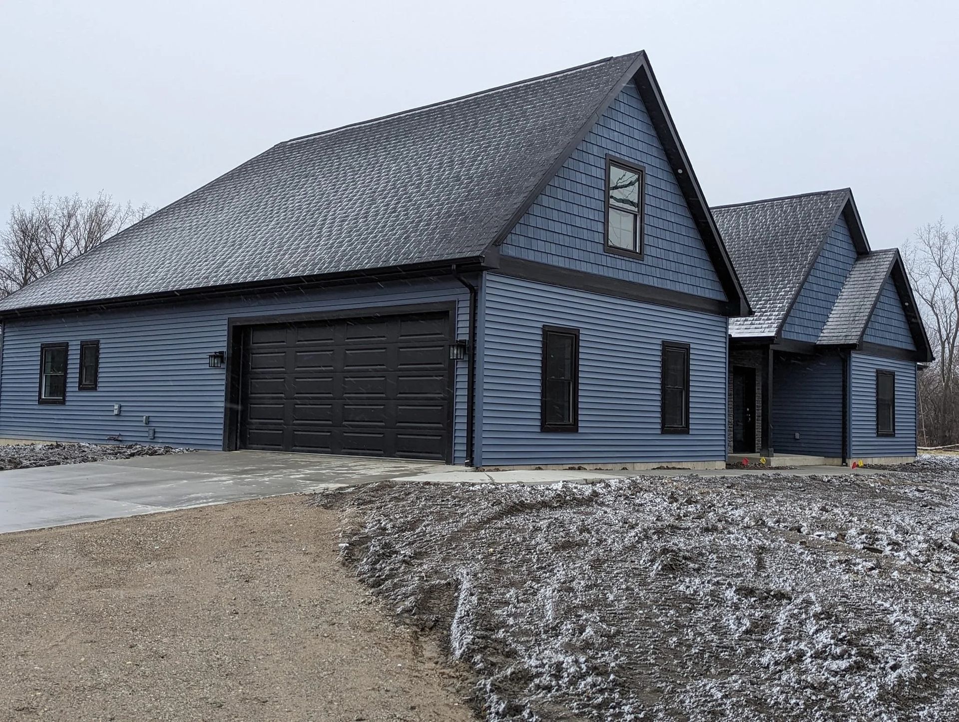Blue farmhouse-style house with attached garage and snowy driveway under an overcast sky