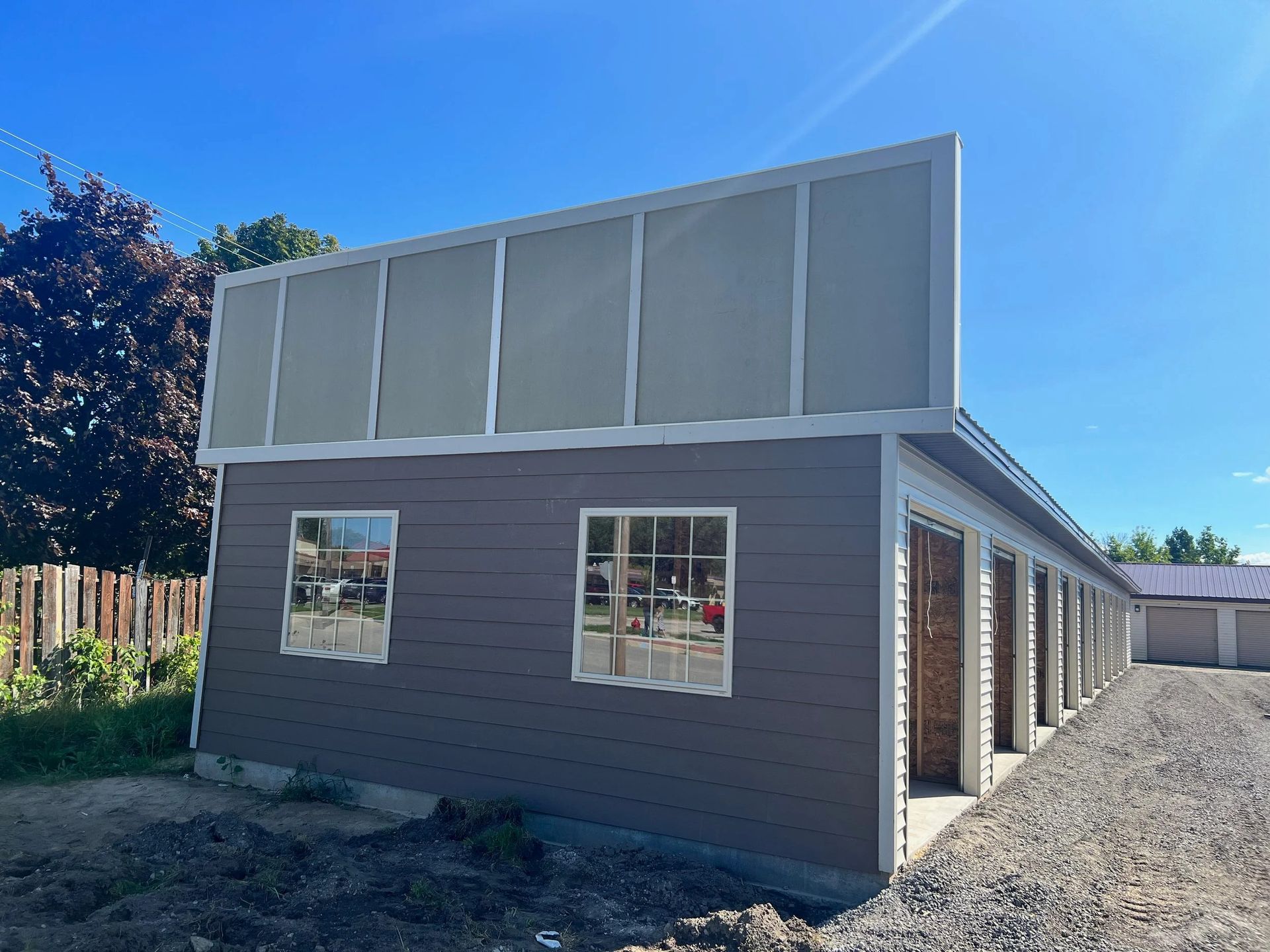 Tan-sided building with large upper windows, white trim, and gravel driveway under a clear blue sky