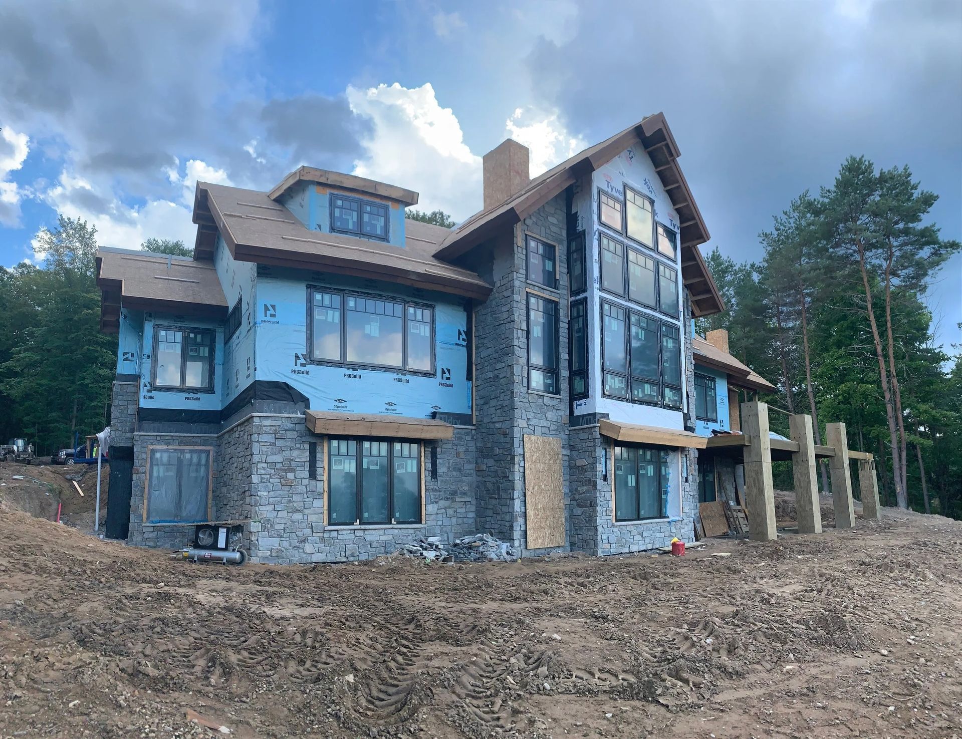 Large modern house under construction with stone and blue siding on a dirt lot under a cloudy sky