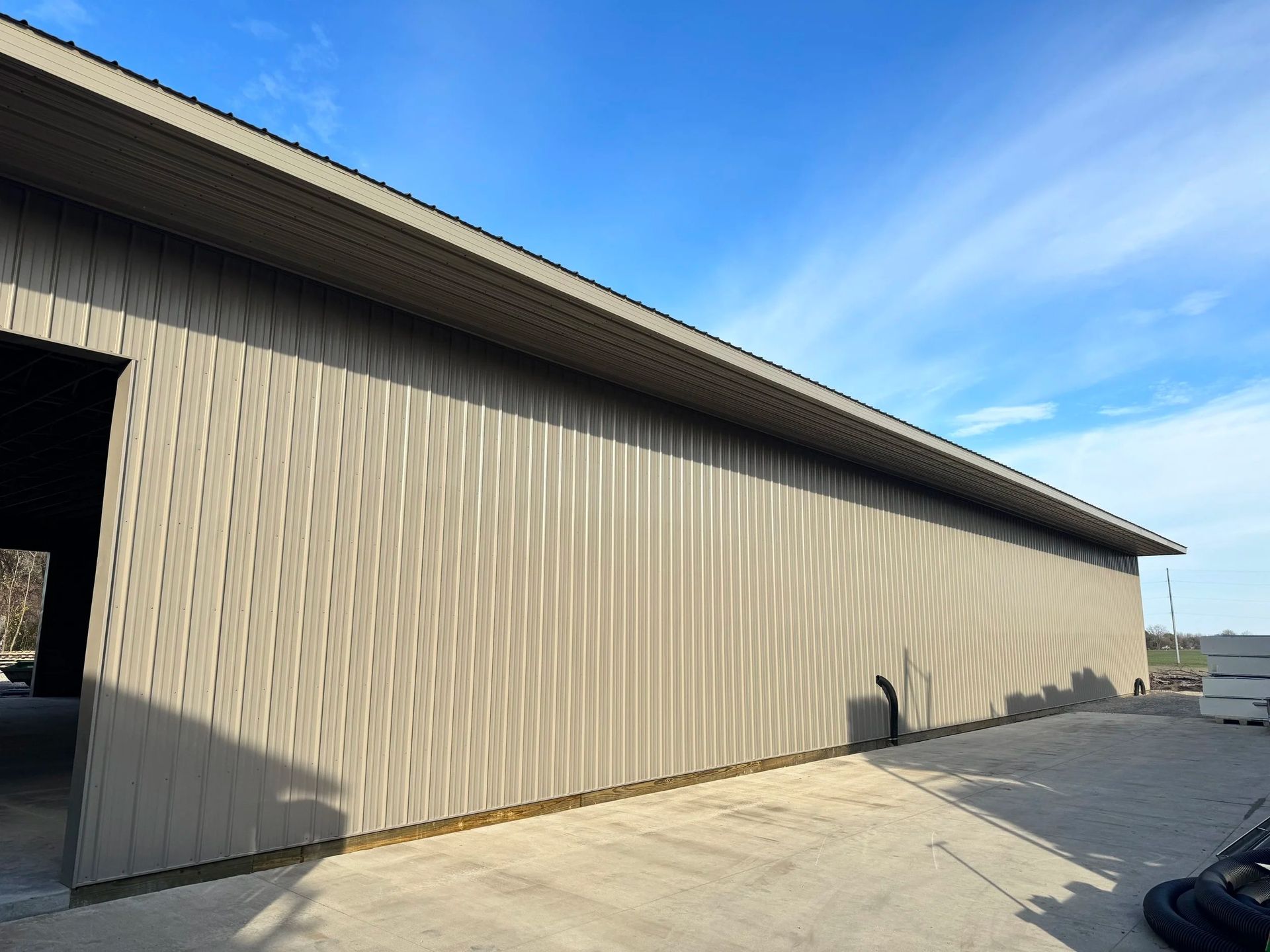 Side view of a beige metal building with a large open doorway and paved lot under a blue sky.