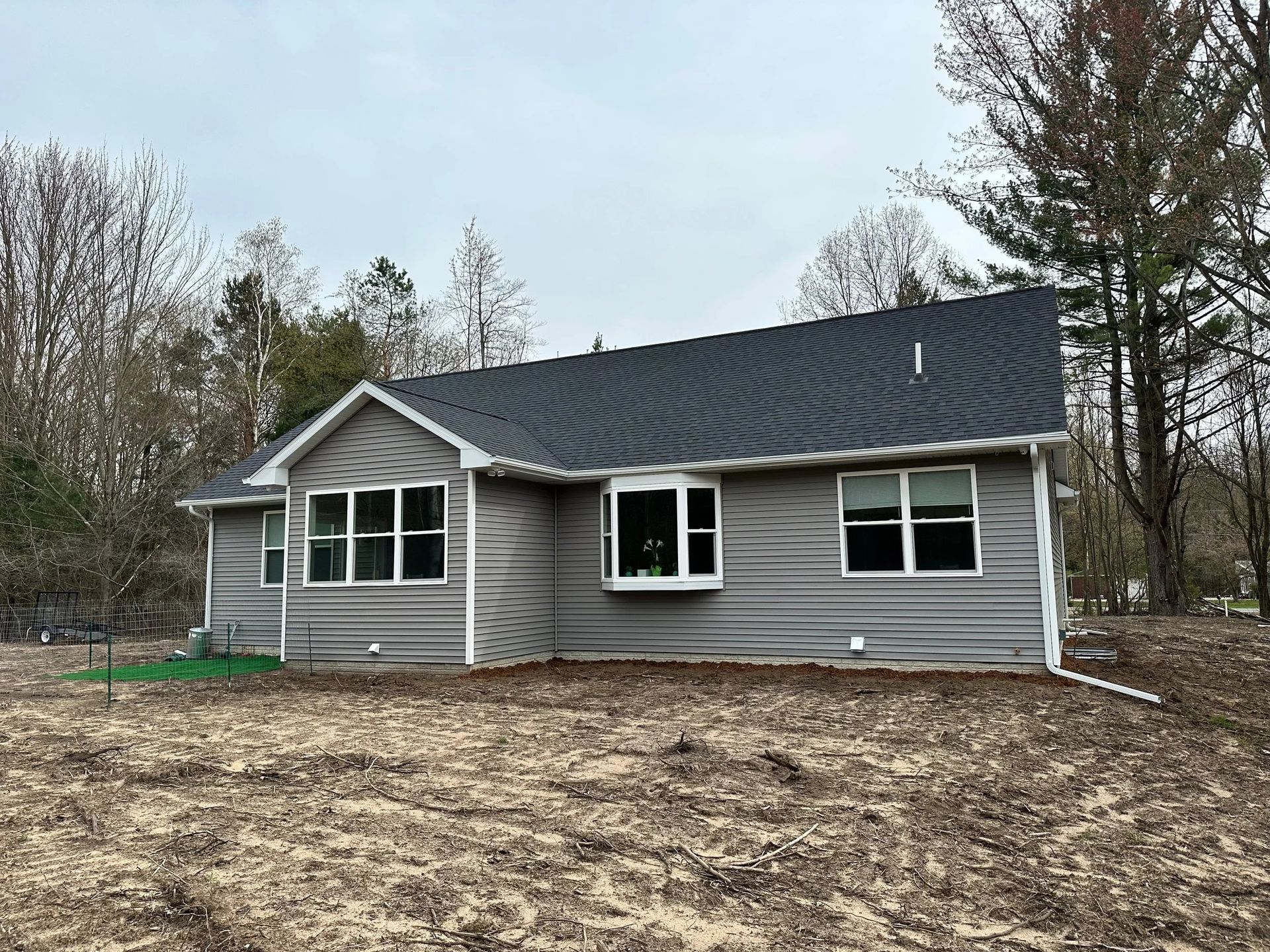 Gray single-story house with black roof in a bare wooded yard