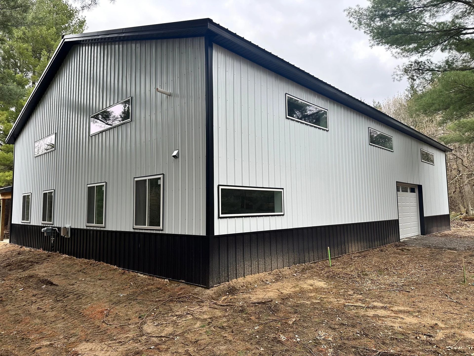 White metal-sided building with black foundation on a wooded hillside, viewed from the corner.