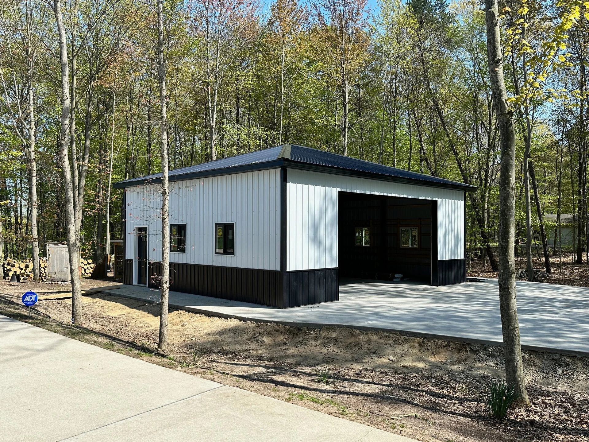 Small white-and-black metal building with a covered porch in a wooded area near a paved path