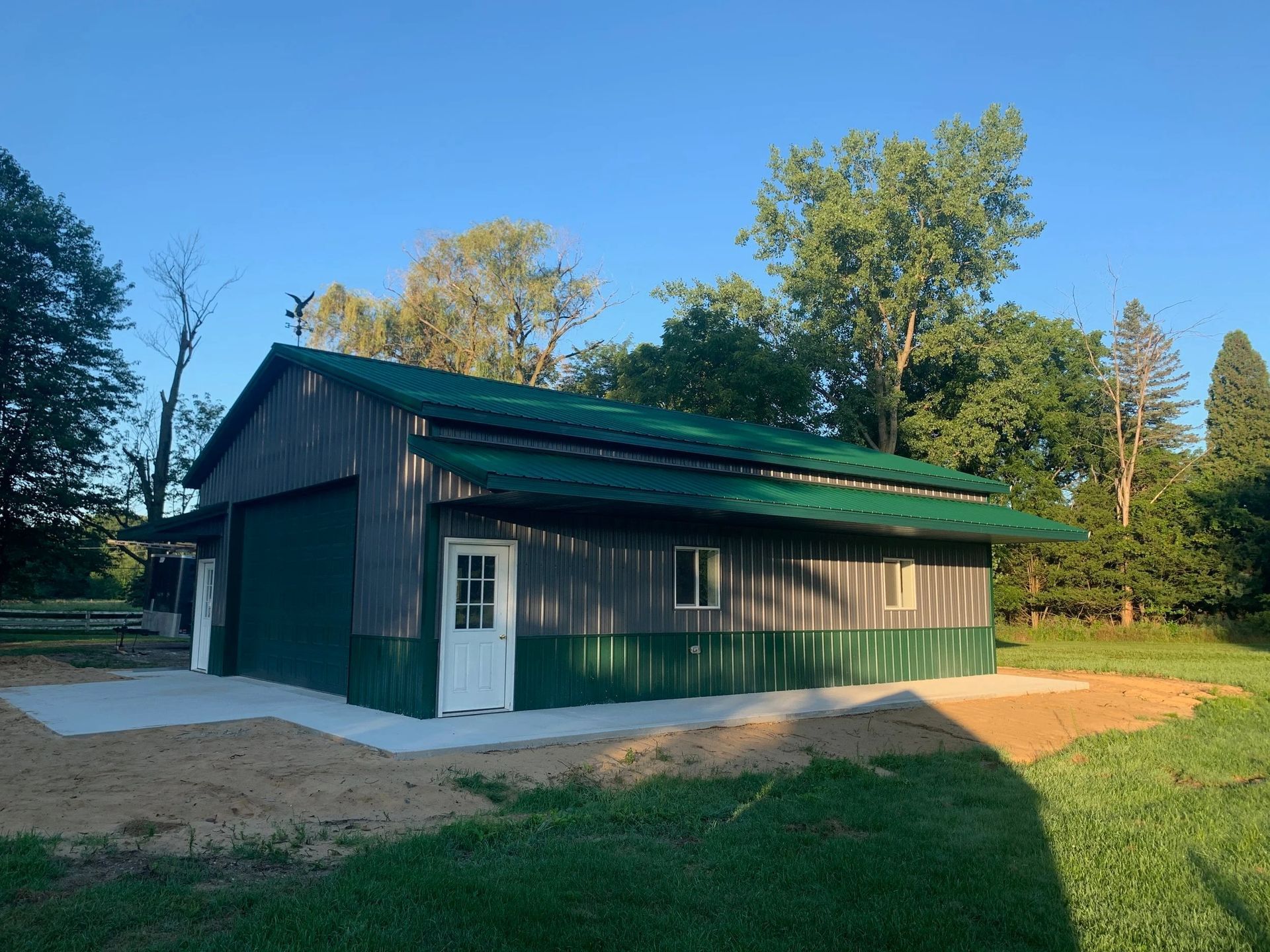 Green metal barn with white door on a dirt lot, surrounded by trees in daylight