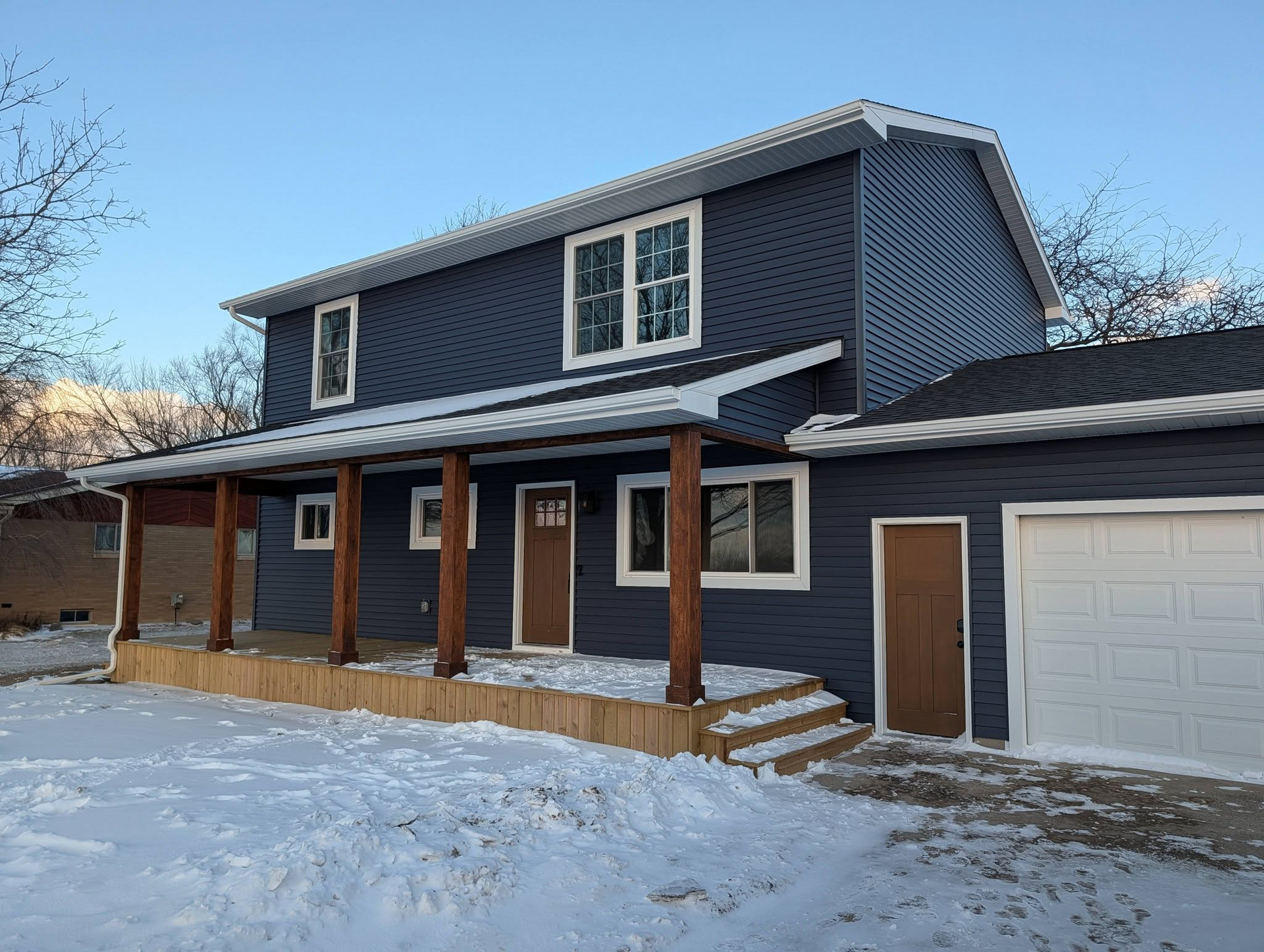 Snow-covered two-story dark blue house with a covered porch and attached garage at dusk