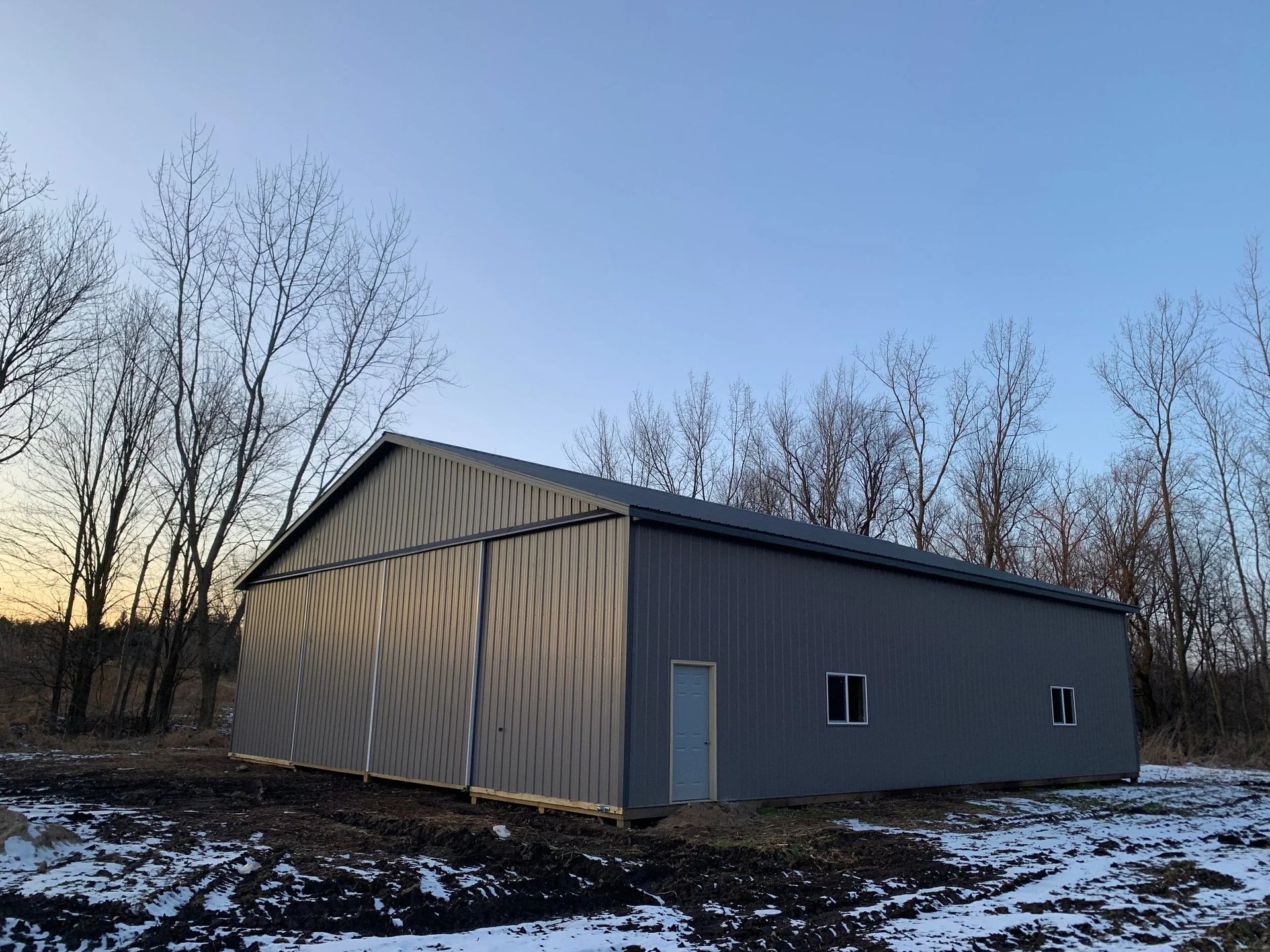 Gray metal barn/shed at dusk with leafless trees and patchy snow around it