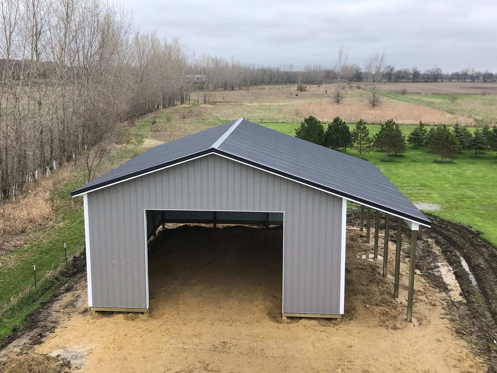 Gray metal barn with open front on a dirt pad beside a green field and trees